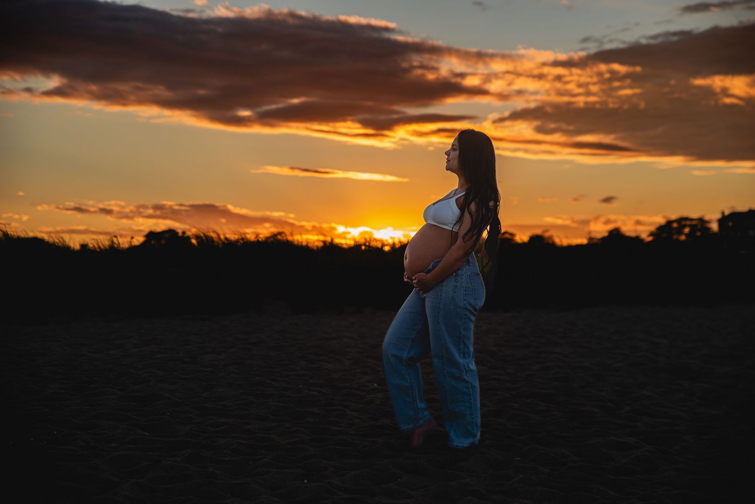 Pregnant woman in white crop top and blue pants stands in open field during golden sunset with dramatic orange sky.