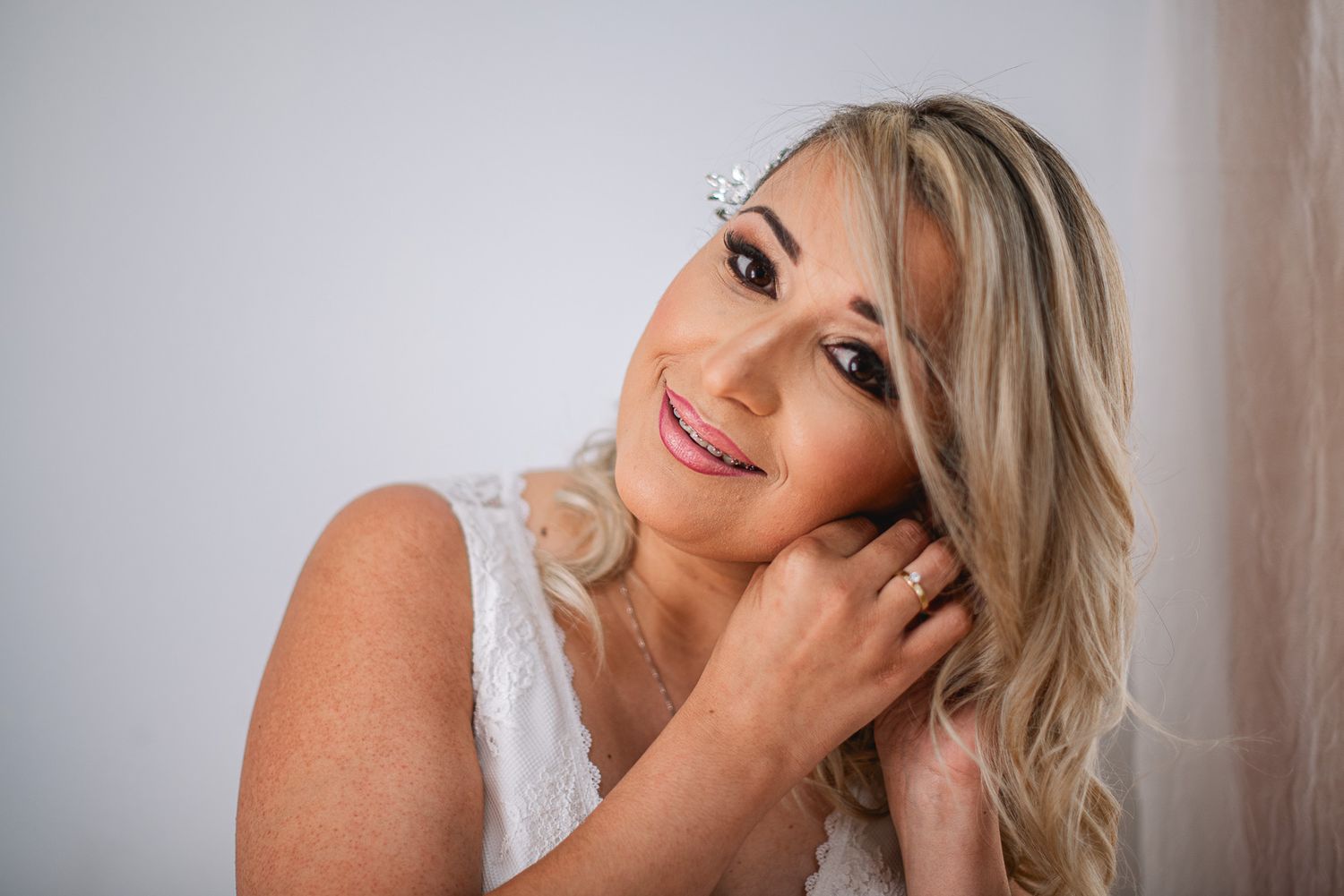 Smiling blonde bride wearing a crystal hair piece and white lace dress, posing elegantly for a wedding portrait.