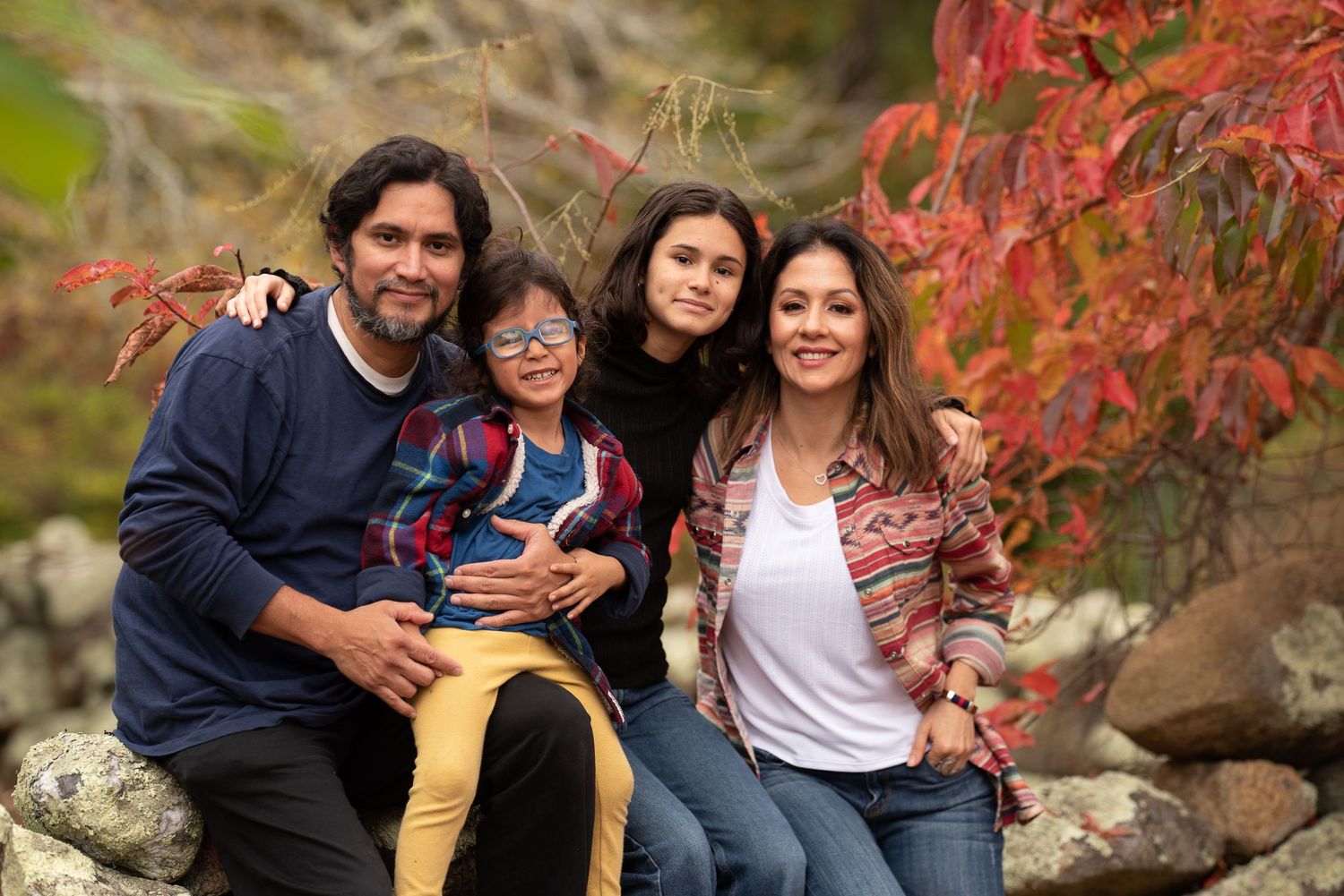 Hispanic family of four smiling together outdoors surrounded by vibrant red autumn foliage on rocks.
