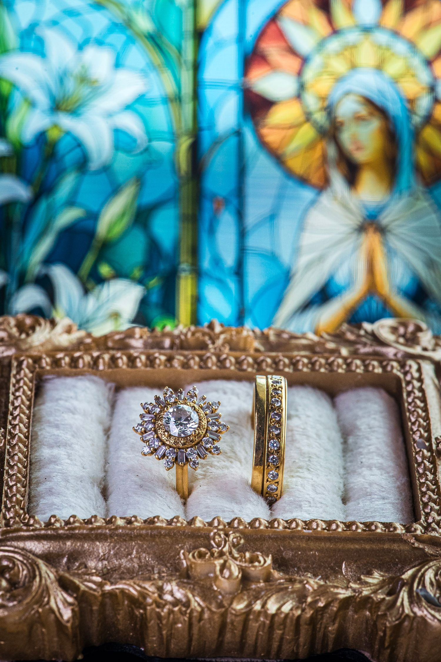 Diamond and gold wedding rings displayed in ornate box before colorful Virgin Mary stained glass.