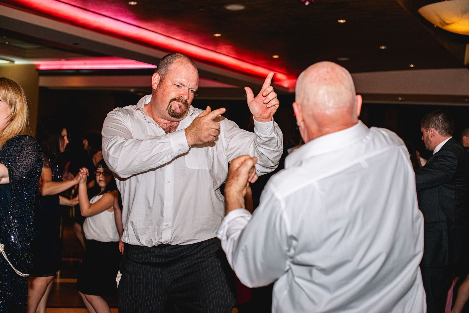 Two bald men dancing energetically at a wedding reception party with guests and red lighting in background.