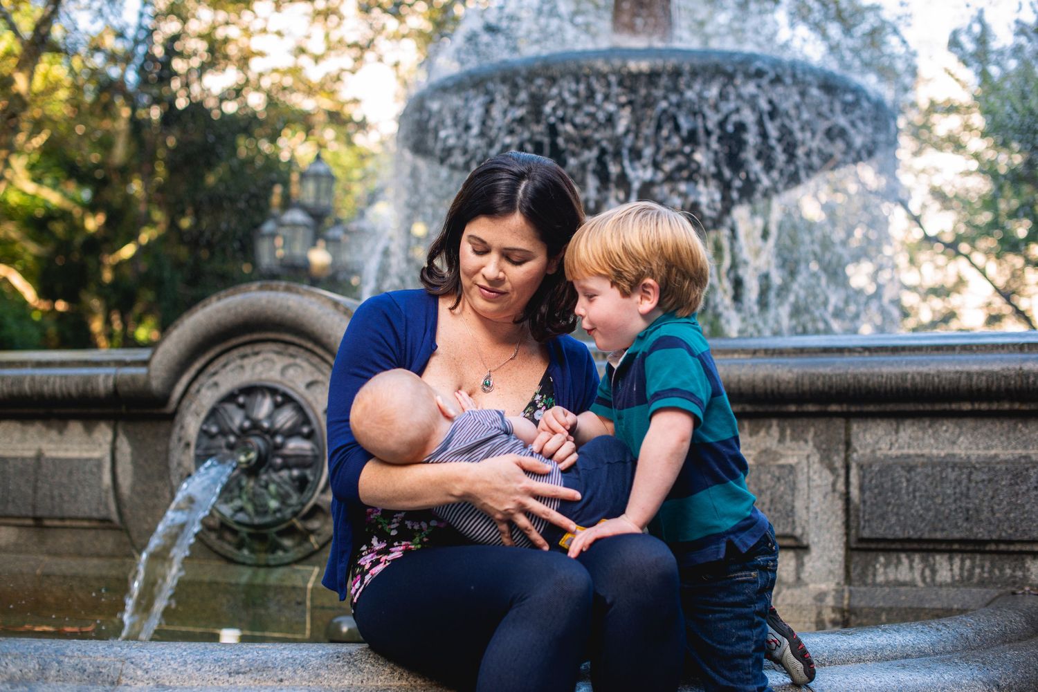 Mother breastfeeding newborn while sitting with young son beside an ornate outdoor fountain.