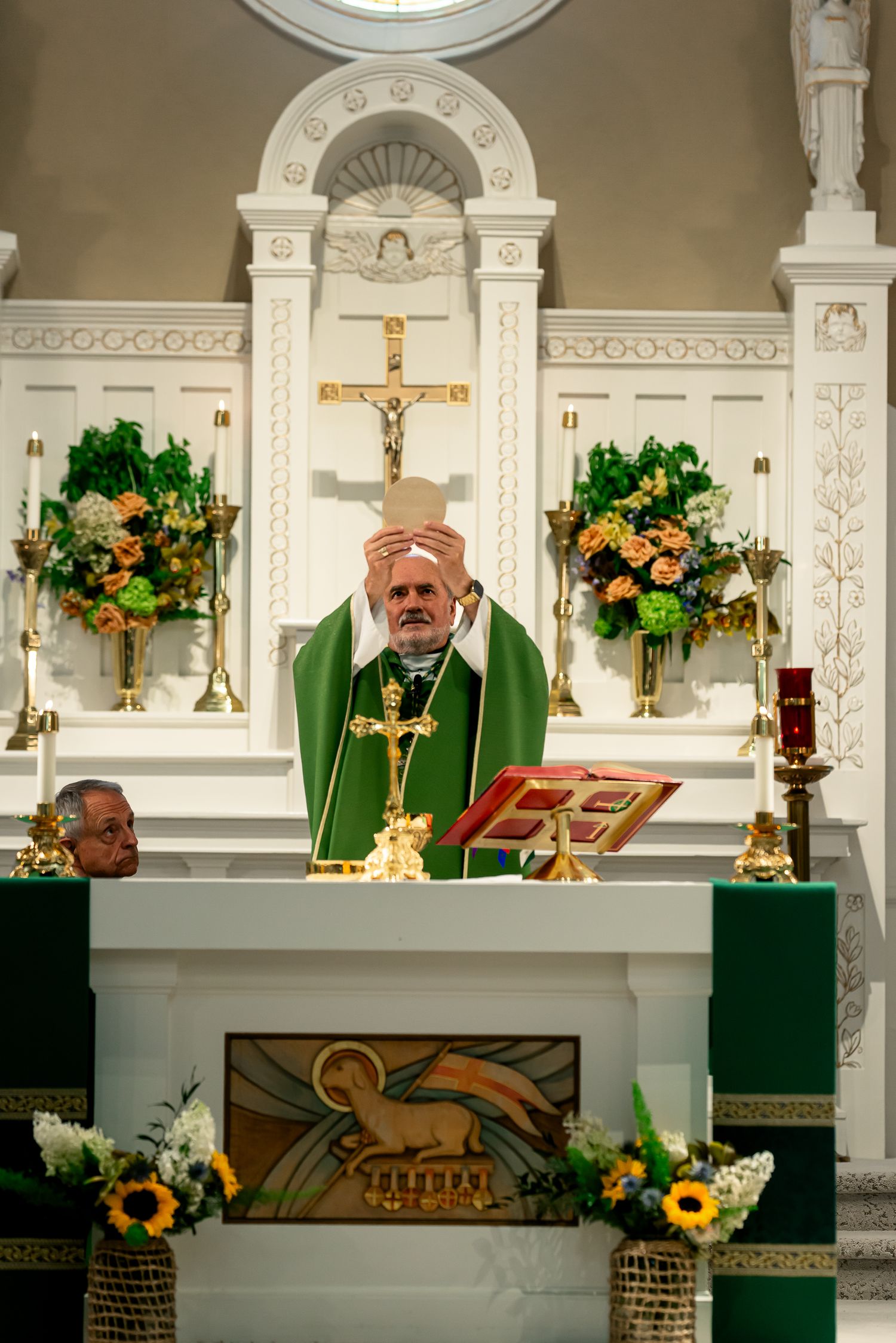 Catholic priest elevating host during Mass at ornate altar with sunflower floral arrangements.