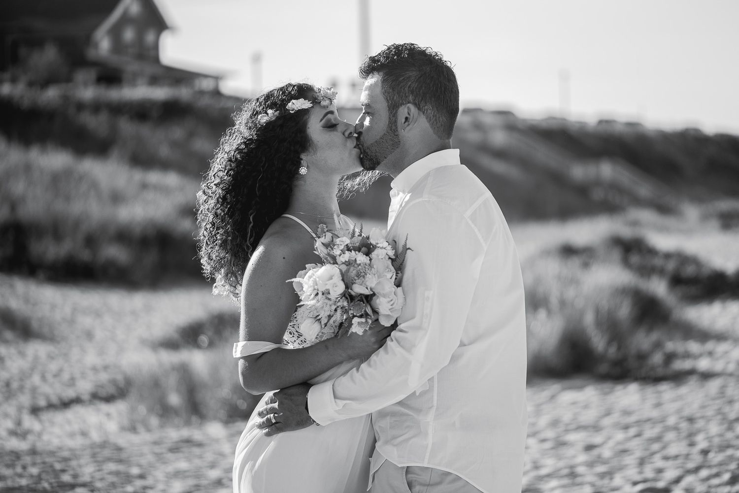 Black and white photo of couple kissing on beach holding bridal bouquet.