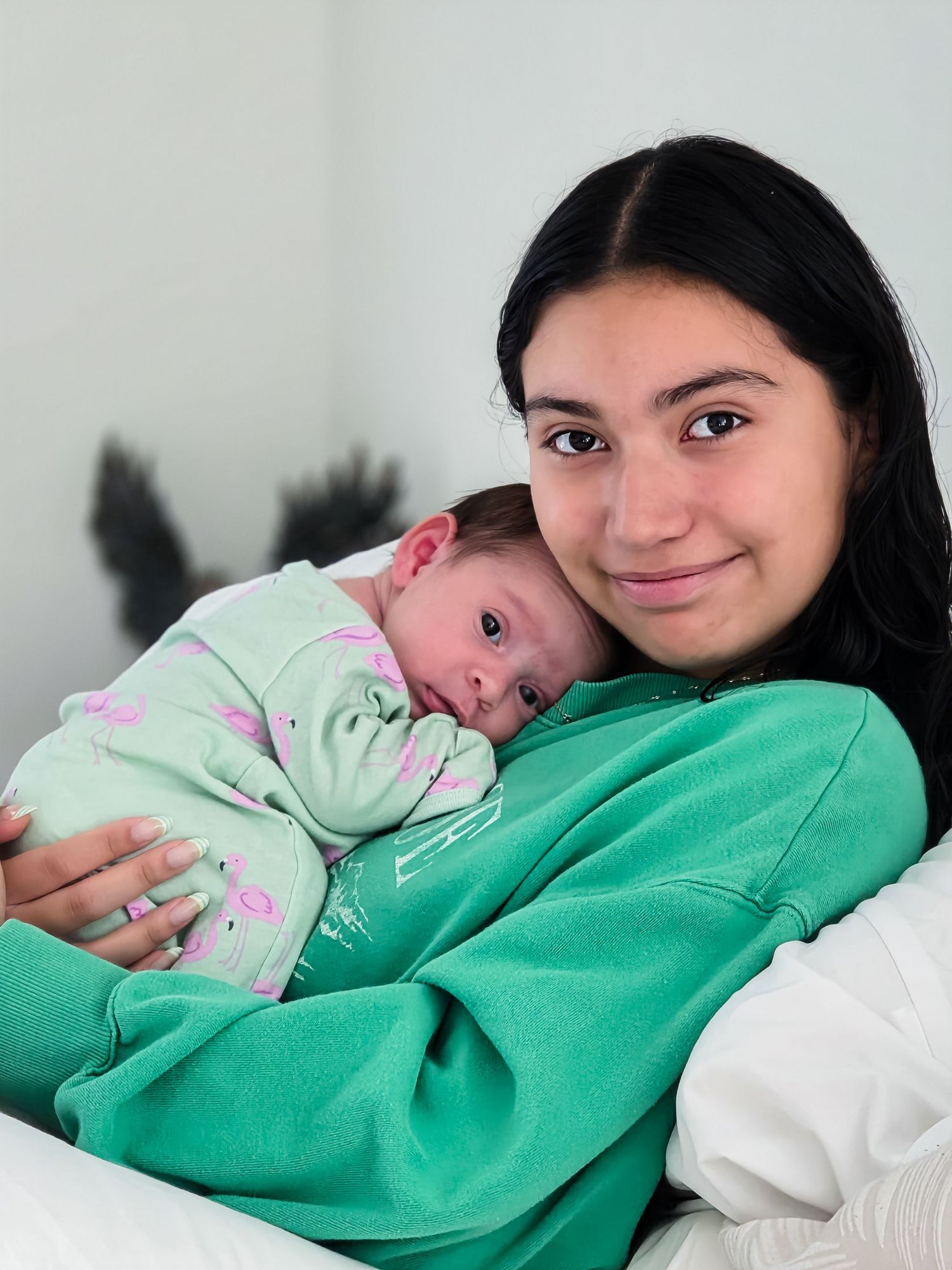 Smiling young woman in a green sweatshirt gently cradles a sleeping newborn baby in a green patterned outfit against her chest.