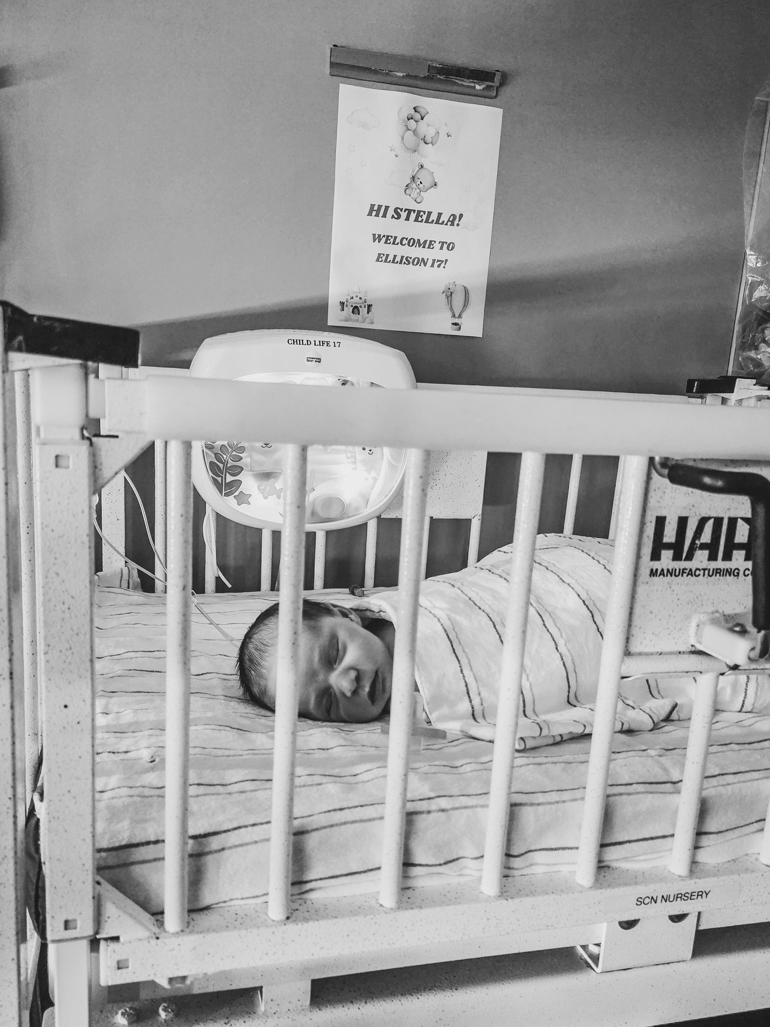 Black and white photo of a swaddled baby sleeping in a hospital crib in the nursery, with a welcome sign for Stella on the wall above.