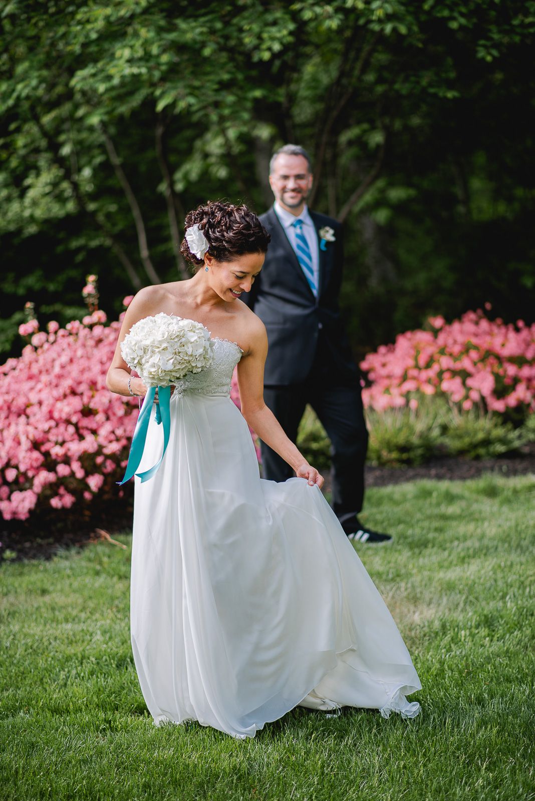 Bride in strapless white gown holding bouquet with teal ribbon, groom in gray suit smiling behind her outdoors.