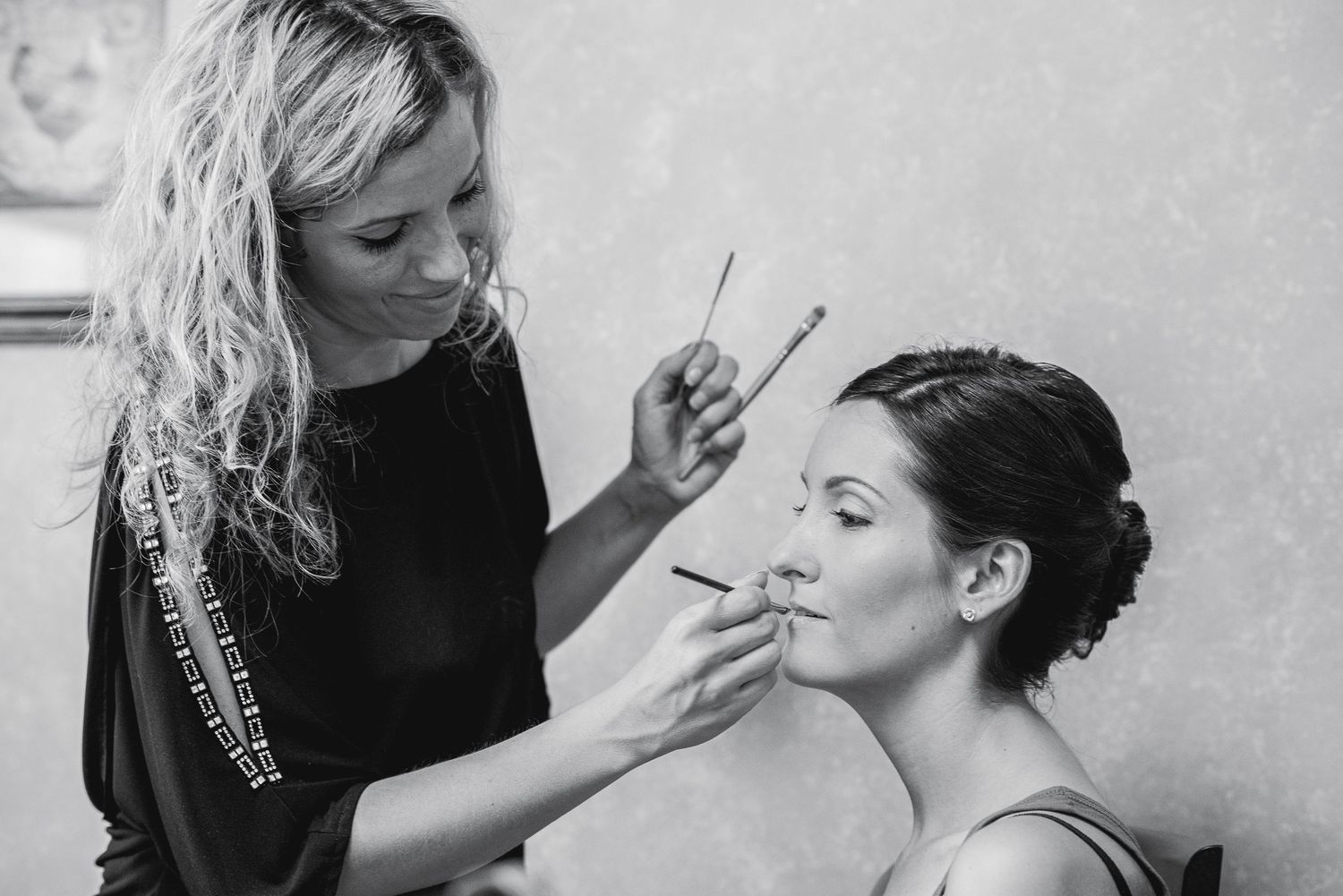 Black and white photo of a makeup artist applying cosmetics to a bride with an updo hairstyle before her wedding.