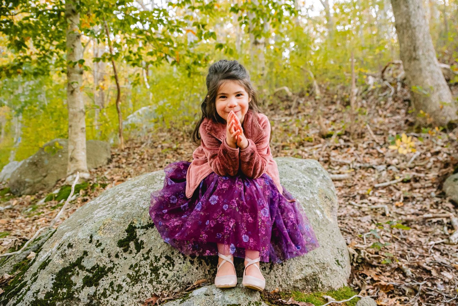 A young girl in a purple tutu skirt sits on a large rock in a forest, smiling and posing for a photo.