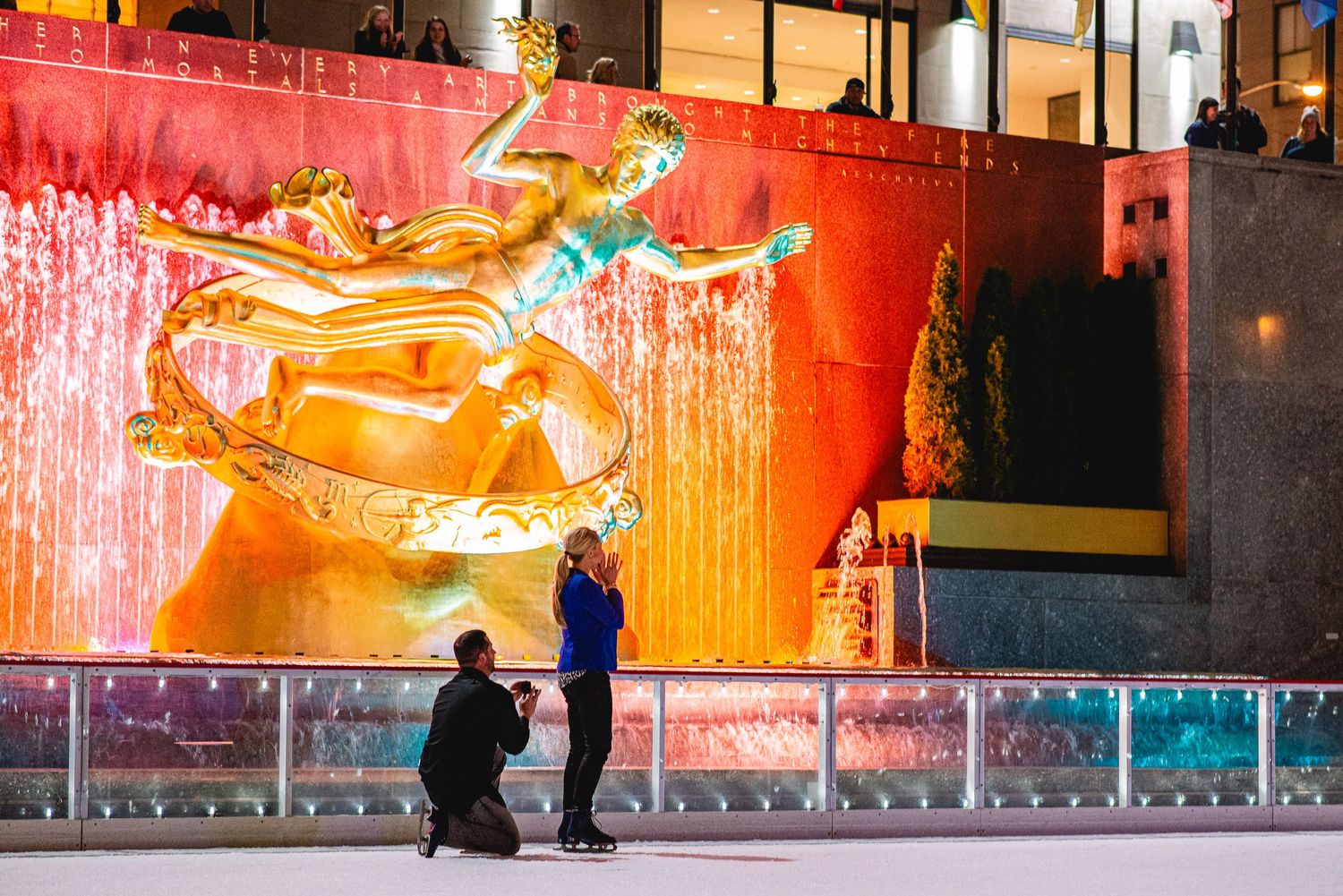 A couple gets engaged on the Rockefeller Center ice rink with the golden Prometheus statue glowing behind them.