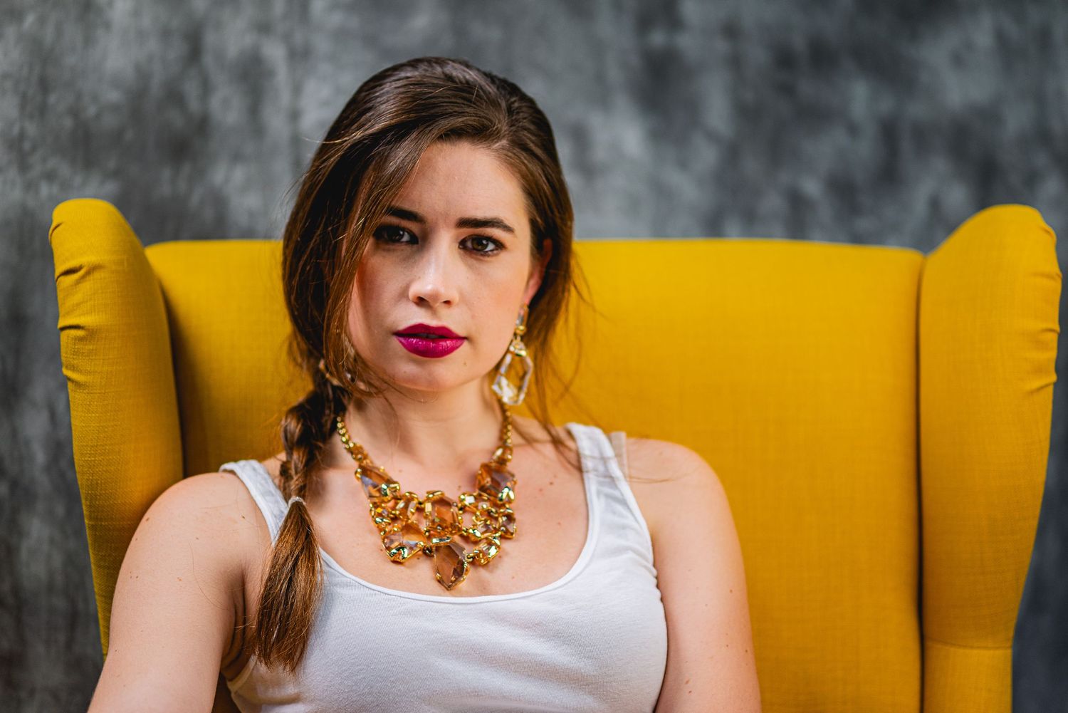 Young woman with braided hair and gold necklace sitting in a yellow armchair, wearing a white top.