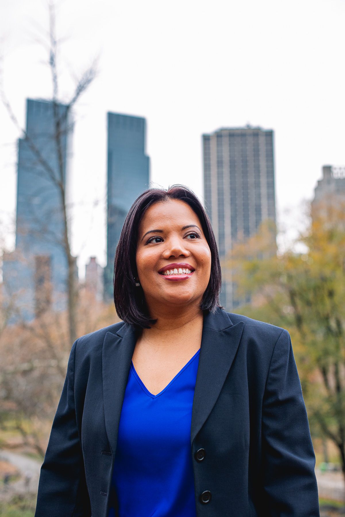Professional woman in blue top and black blazer smiling outdoors with NYC skyline in background.