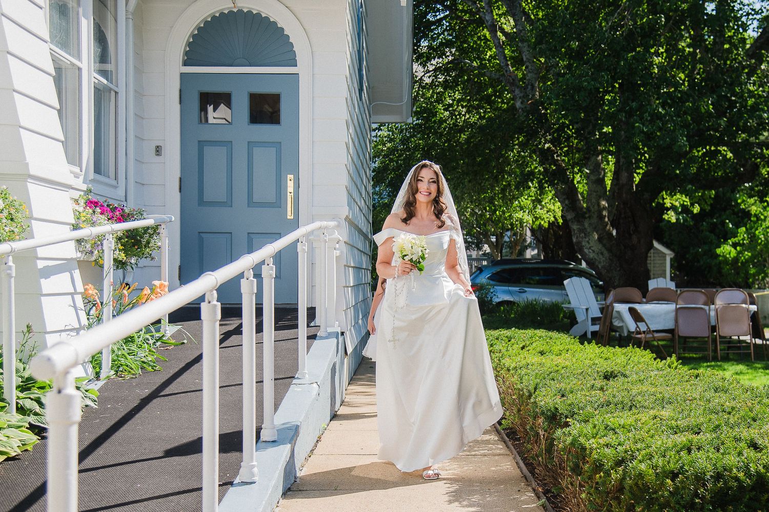 Bride in white gown holding bouquet walks along garden path toward blue door of elegant venue on sunny day.
