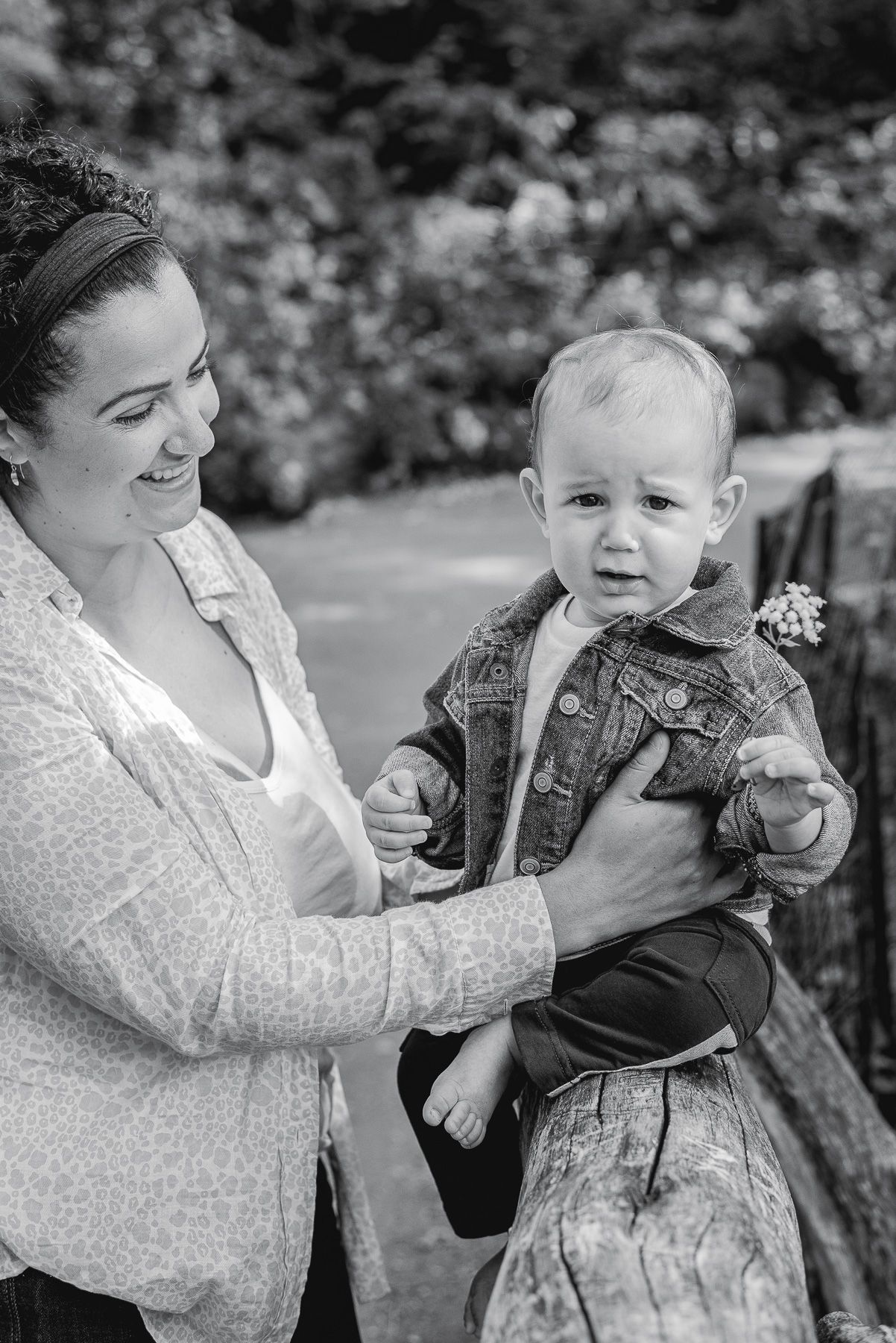 Black and white photo of a smiling woman holding a cute baby in a denim jacket outdoors.