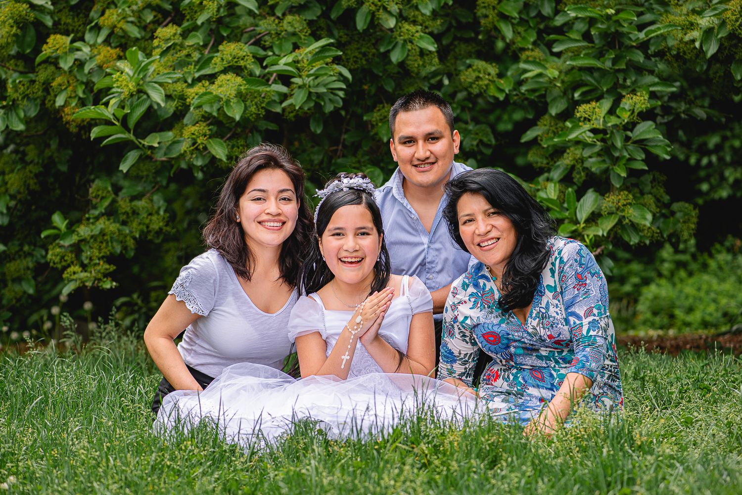 Happy family of four smiling together on green grass outdoors, wearing white and floral outfits in a lush garden setting.