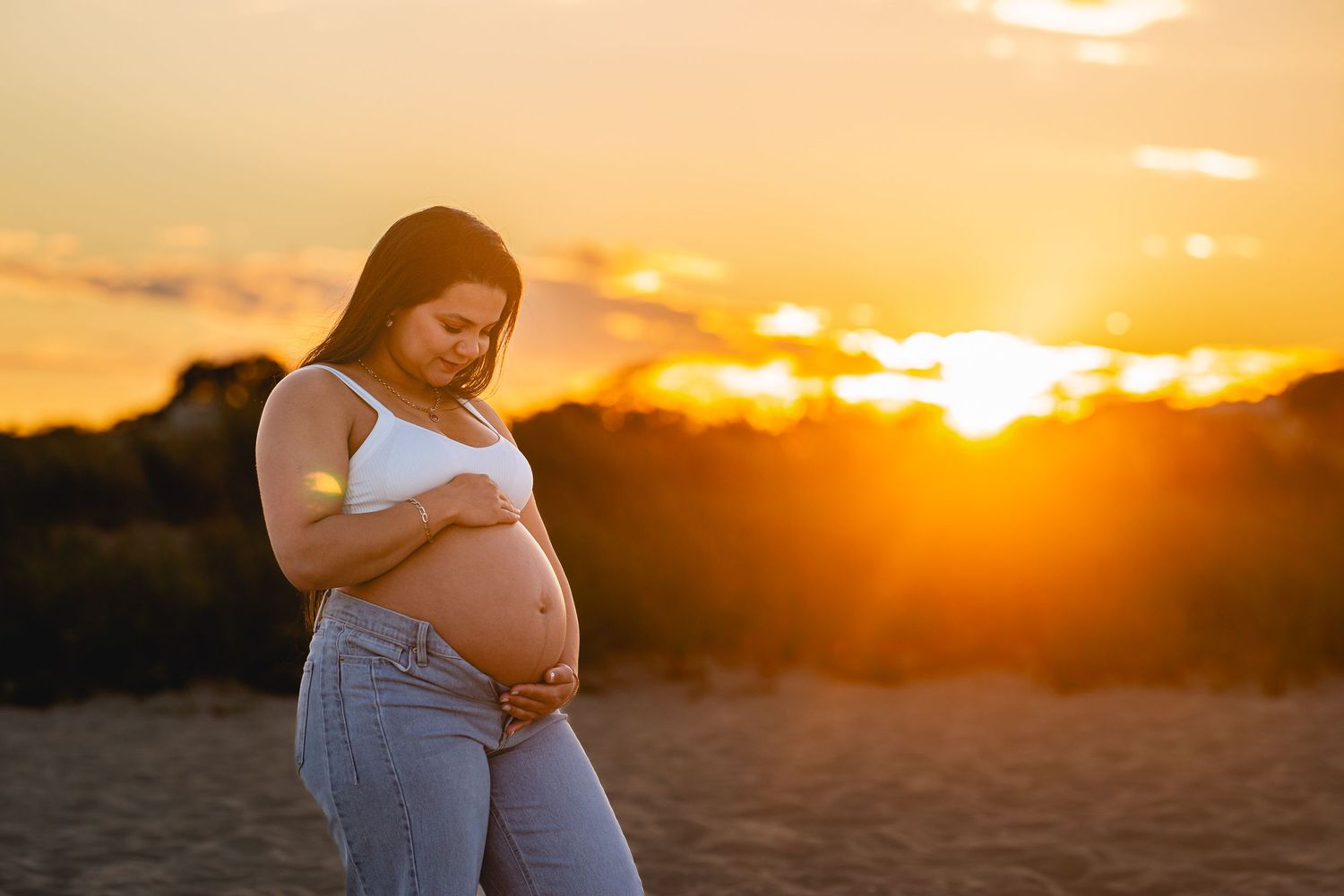 Pregnant woman cradling her baby bump at golden sunset on a beach, wearing white crop top and jeans.
