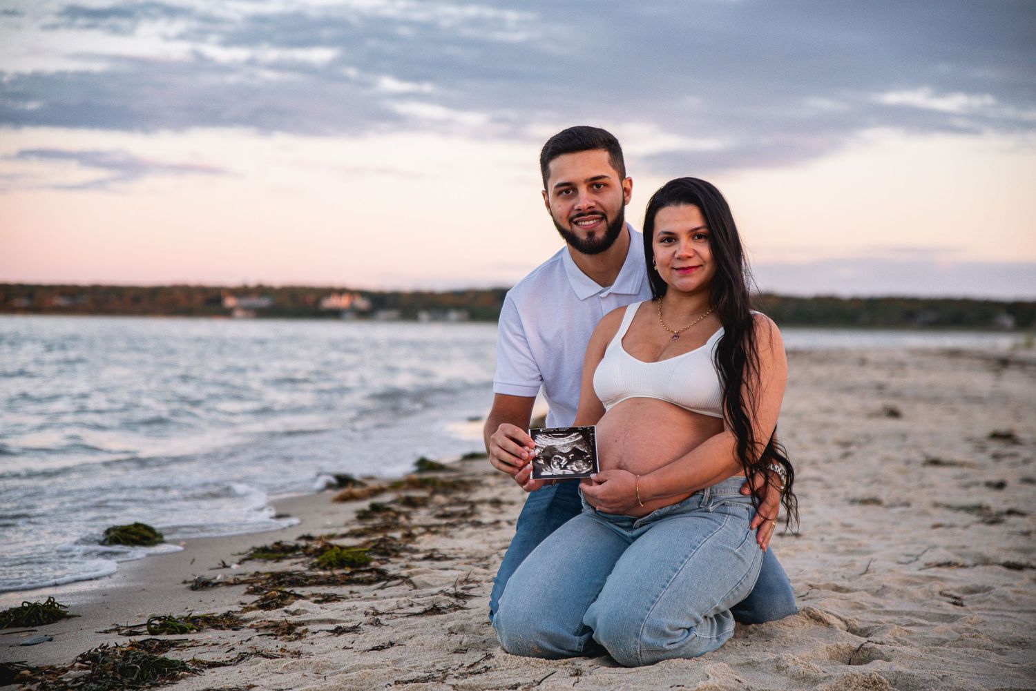 Couple kneeling on beach at sunset, woman showing pregnant belly and ultrasound photo, romantic maternity photoshoot.