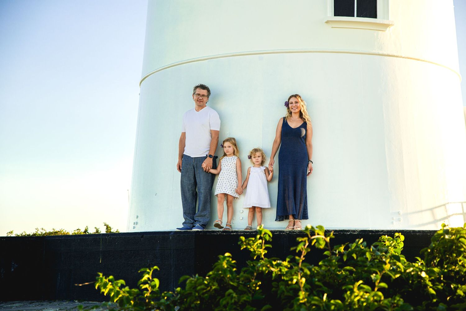 Family of four posing in front of a white lighthouse surrounded by green shrubs on a sunny day.