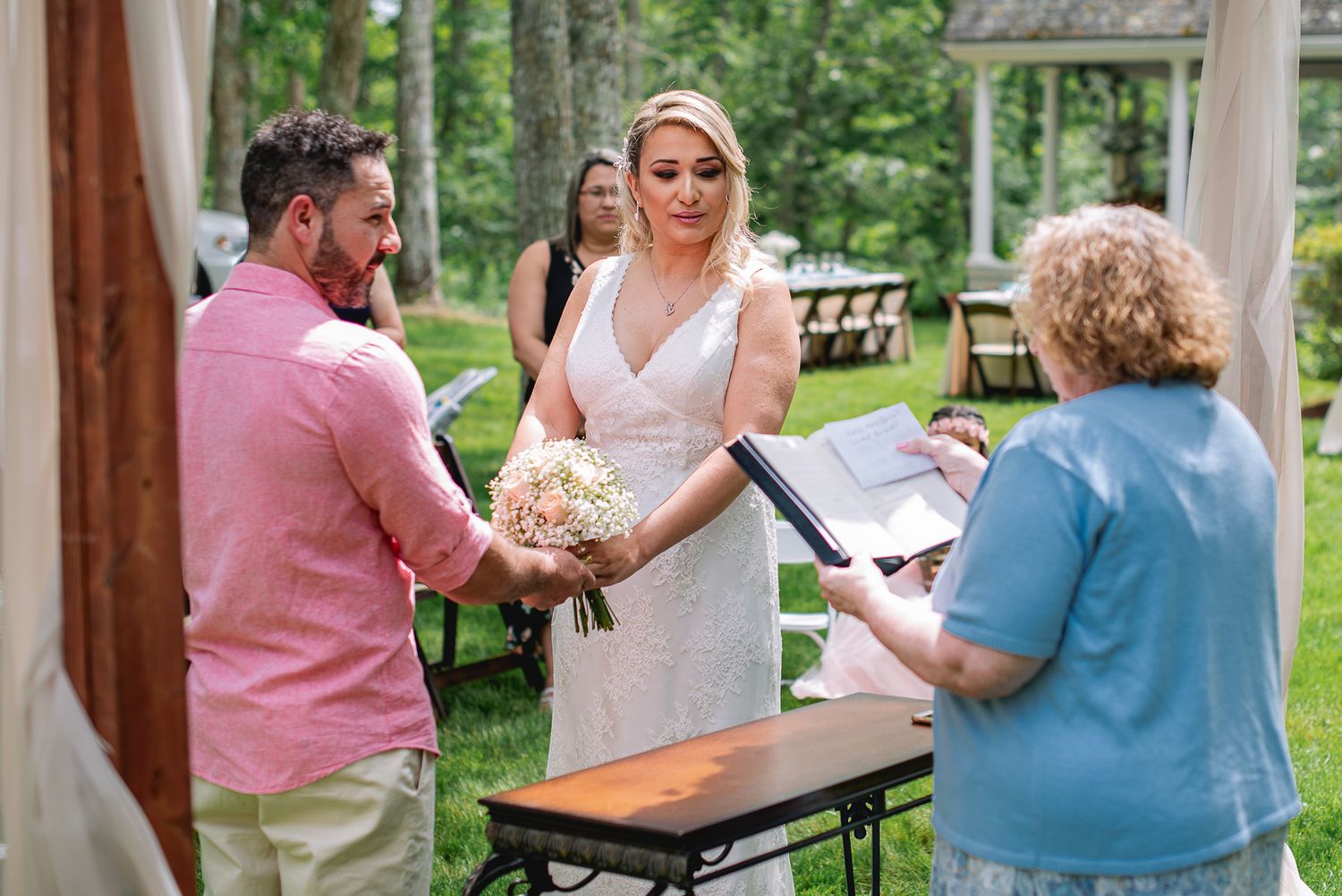 Outdoor wedding ceremony with bride holding bouquet, groom in pink shirt, and officiant reading vows in a garden setting.