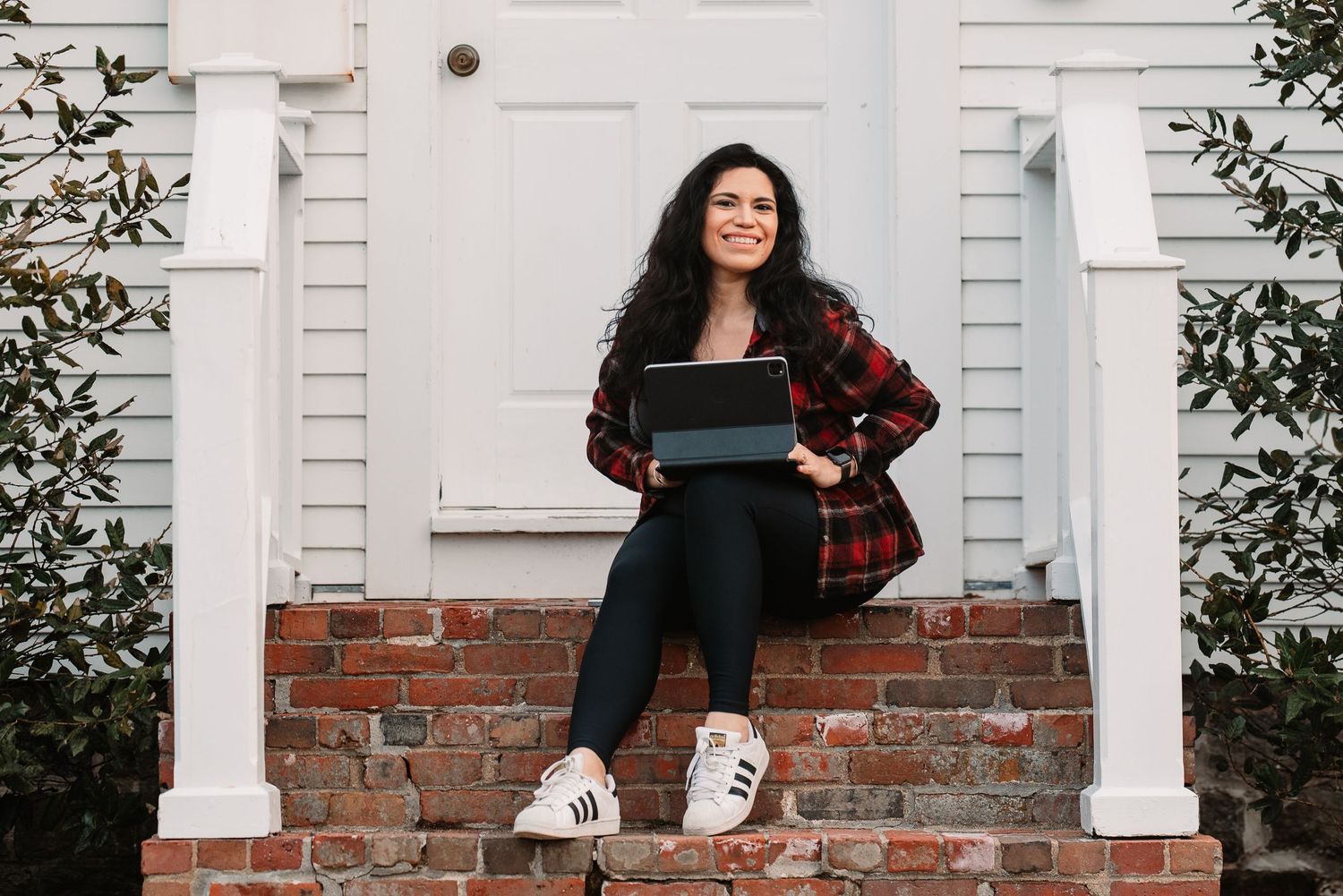 Woman in plaid shirt sitting on brick steps with laptop, smiling outdoors.