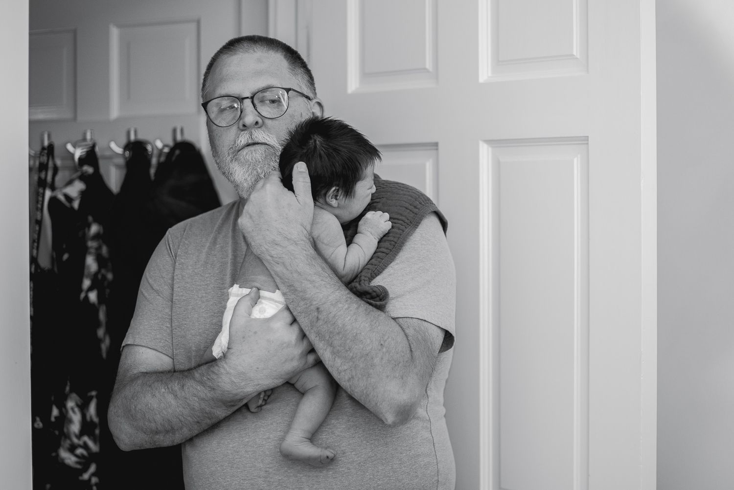 Black and white photo of an older man with glasses holding a newborn baby near a closet.