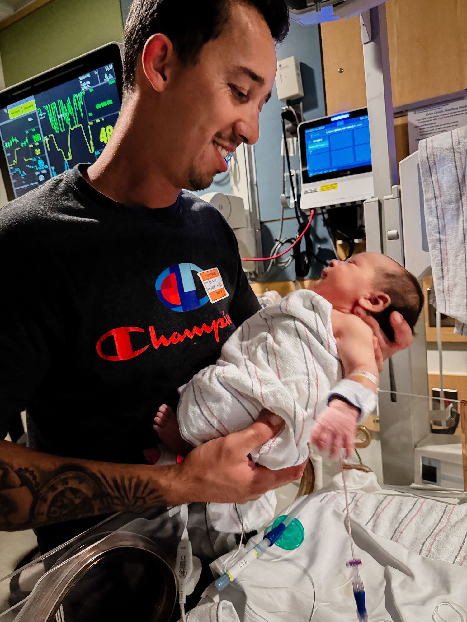 Smiling man in a Champion shirt gently holds a swaddled newborn baby in a hospital room with monitors and medical equipment nearby.
