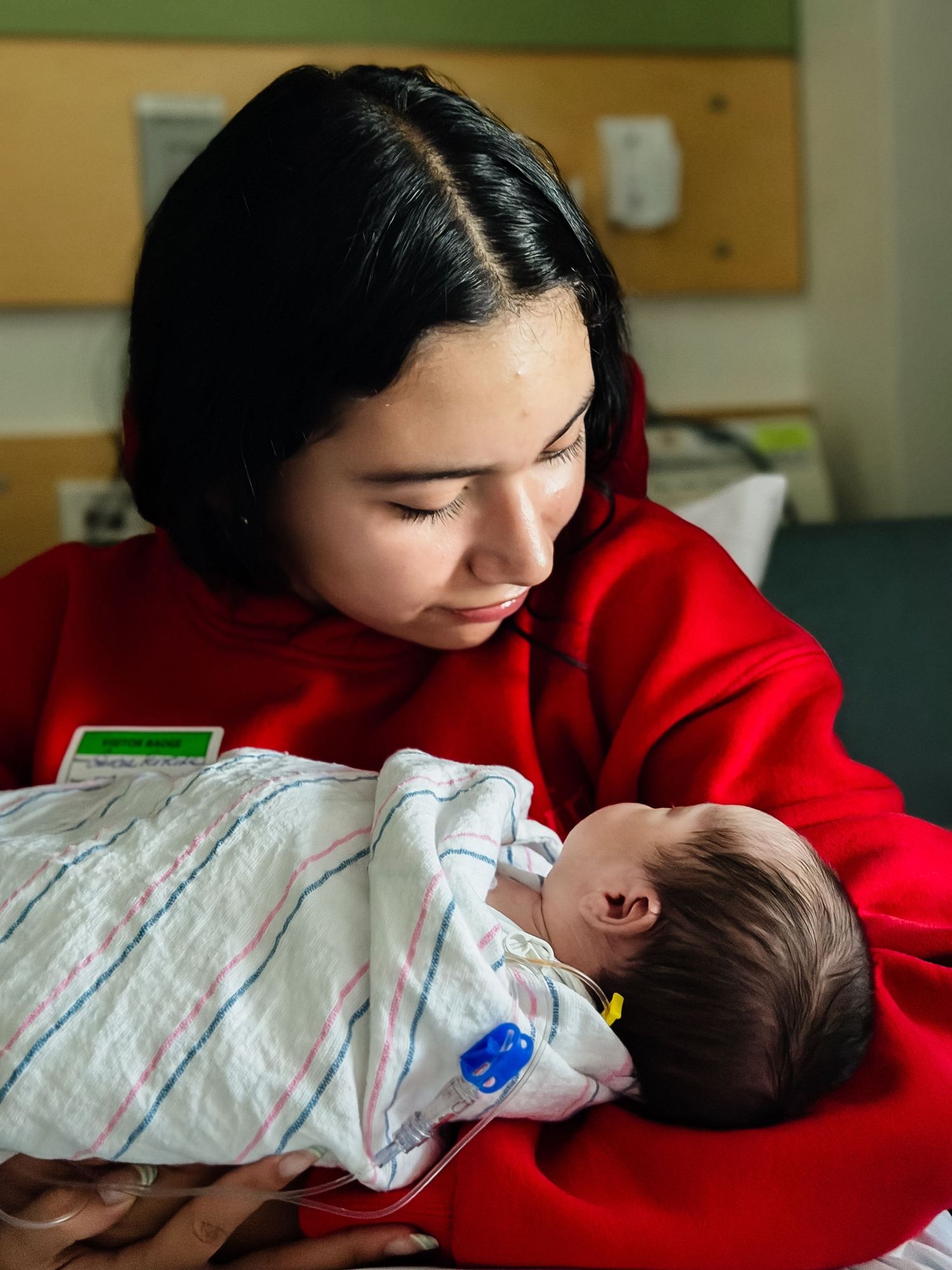 Young mother in a red sweater gently cradles a sleeping newborn wrapped in a striped hospital blanket in a hospital room.