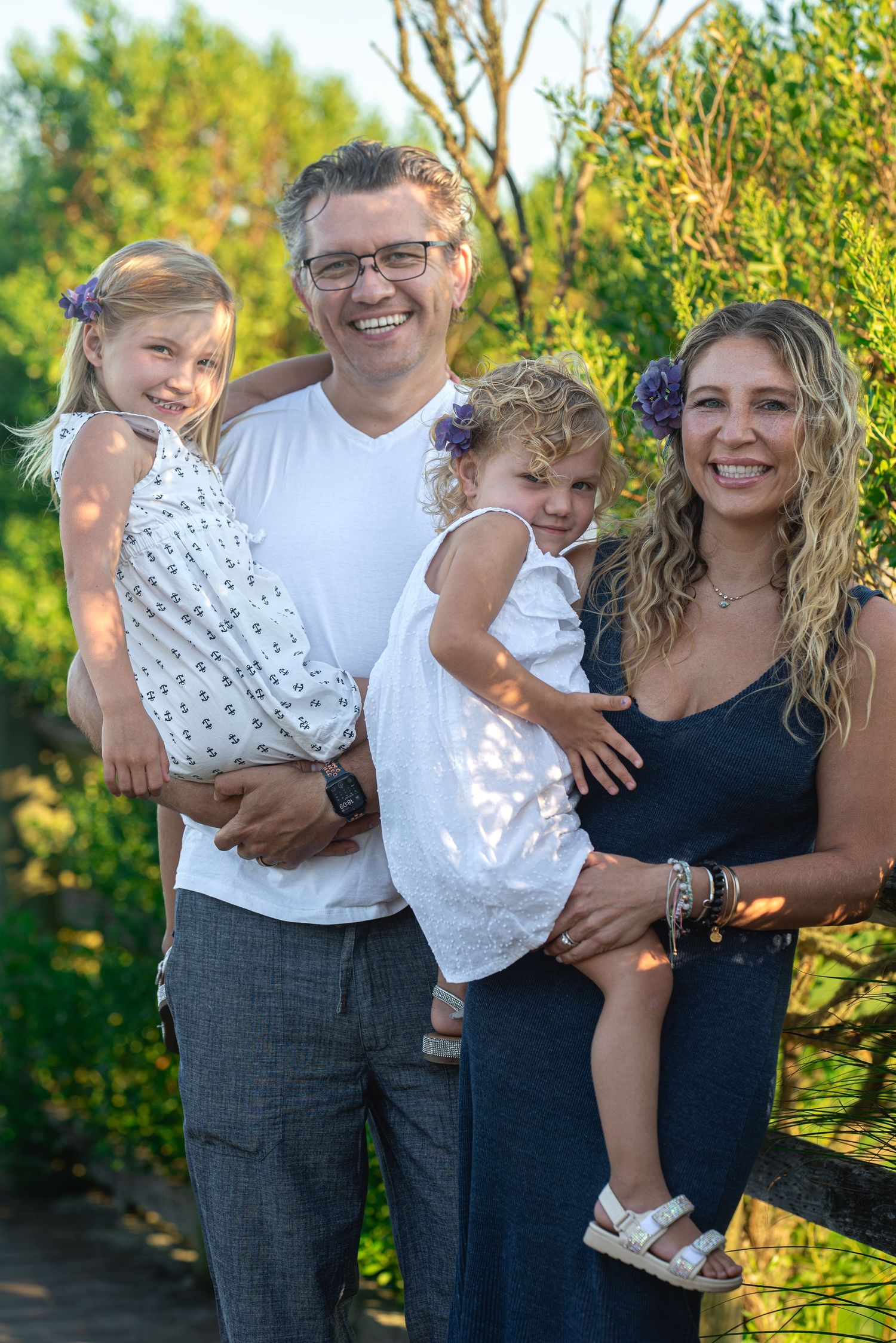 Happy family of four posing outdoors, parents holding two young blonde girls, smiling in a lush green garden setting.