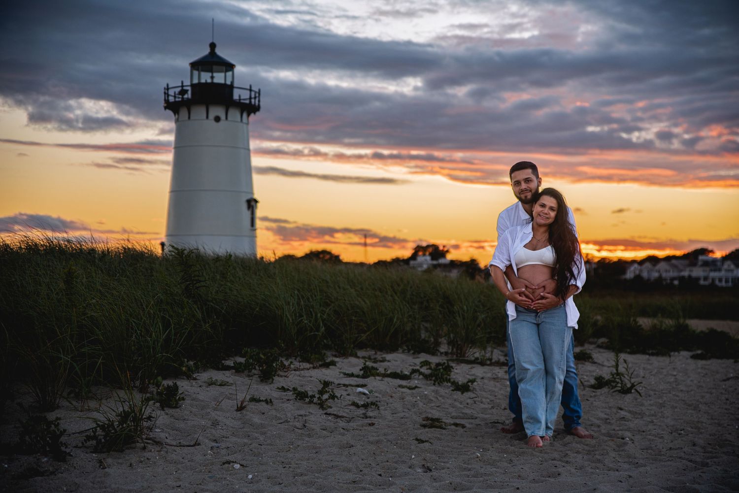 Couple embracing at sunset near a white lighthouse on a sandy beach with dramatic cloudy sky.
