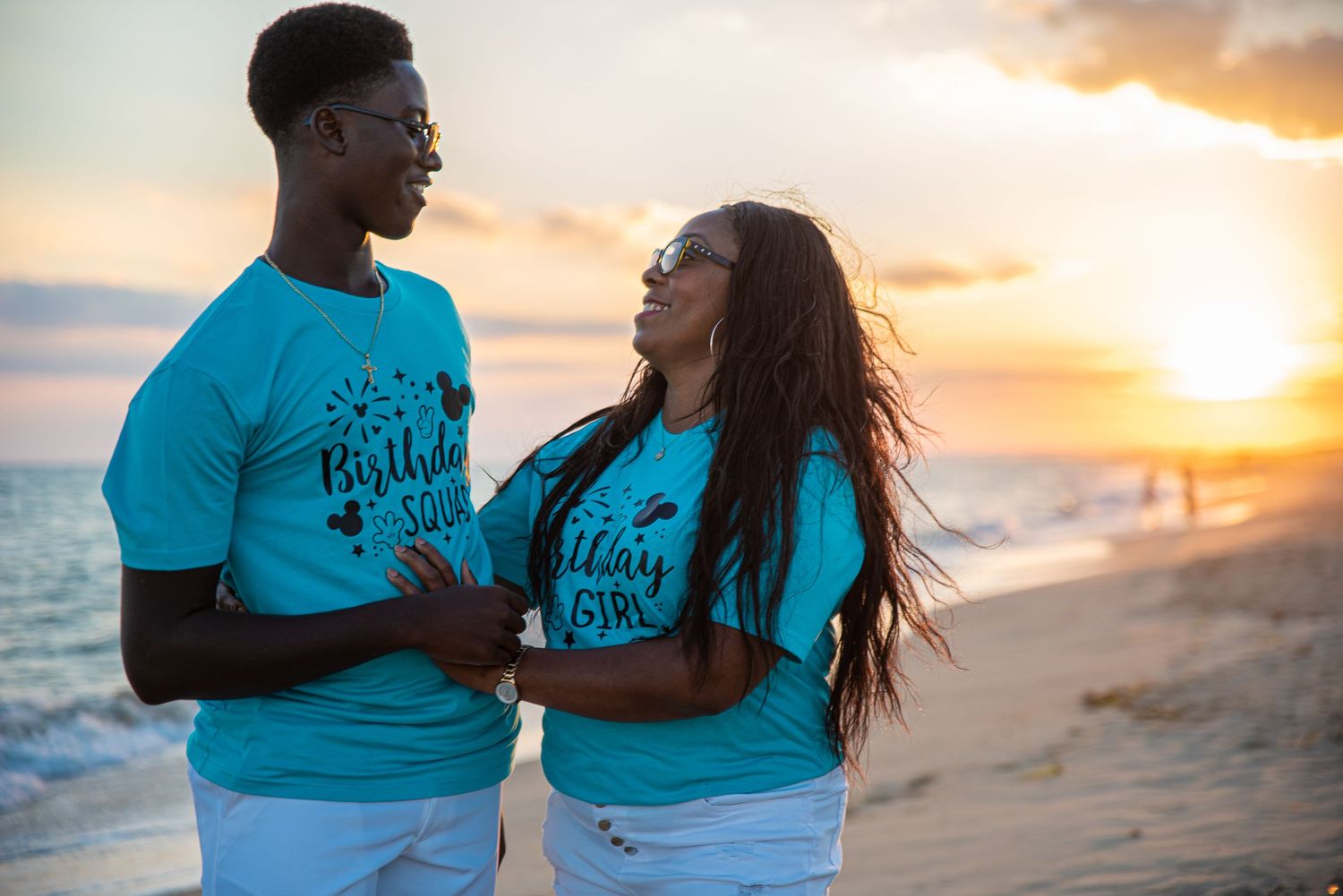Black couple in matching teal birthday shirts smiling at each other on a beach at sunset.