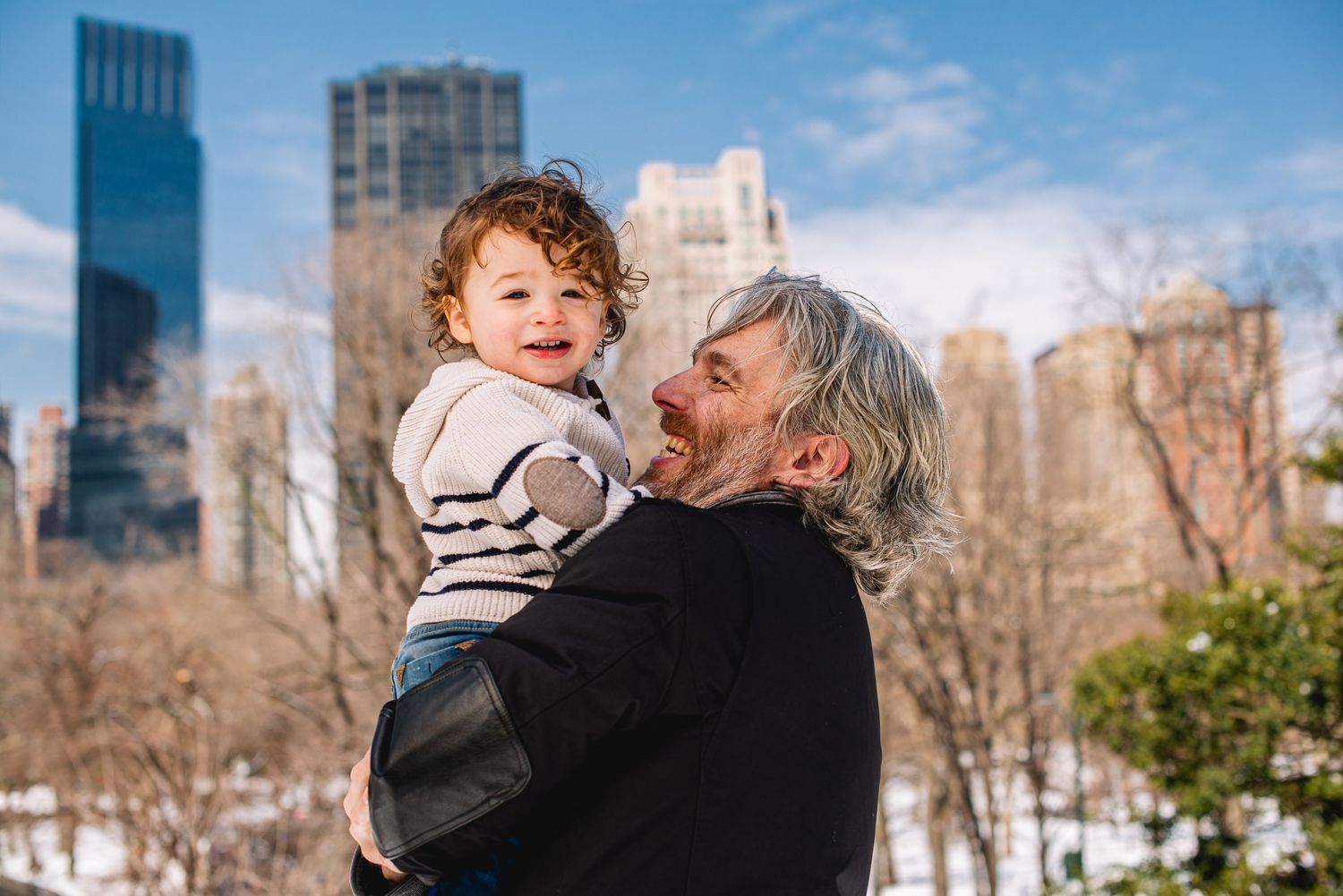 Man with gray hair holds laughing toddler in Central Park with NYC skyline in winter background.