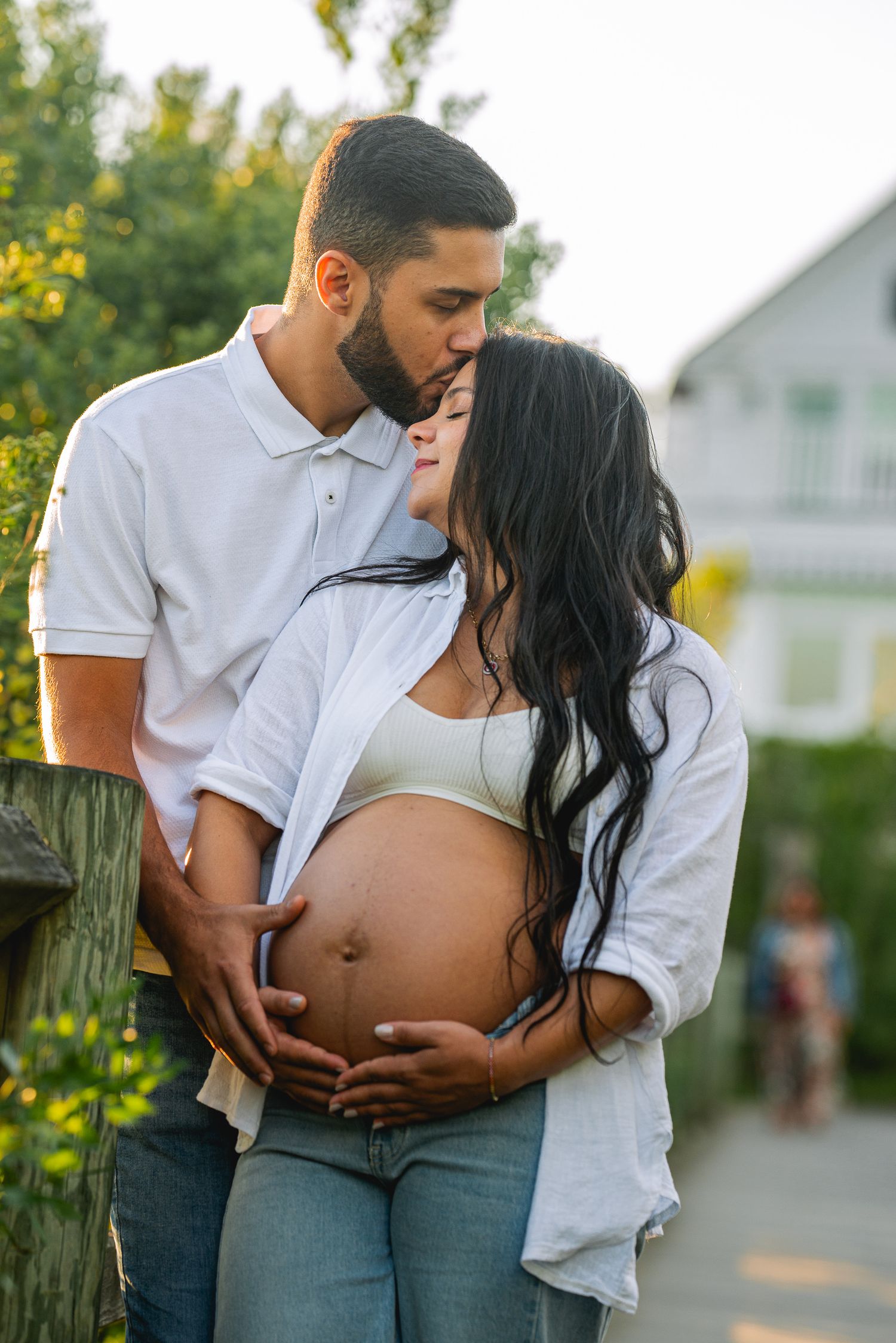 A man kisses a pregnant woman's head while holding her belly outdoors in a tender maternity photo.