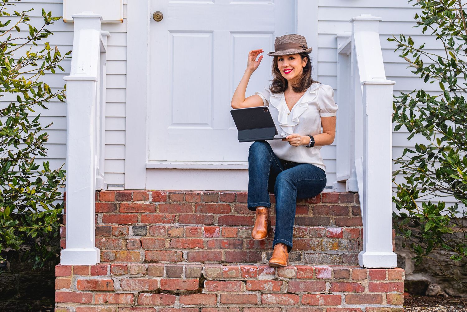 Woman in hat sits on brick steps with laptop outside a charming white house.