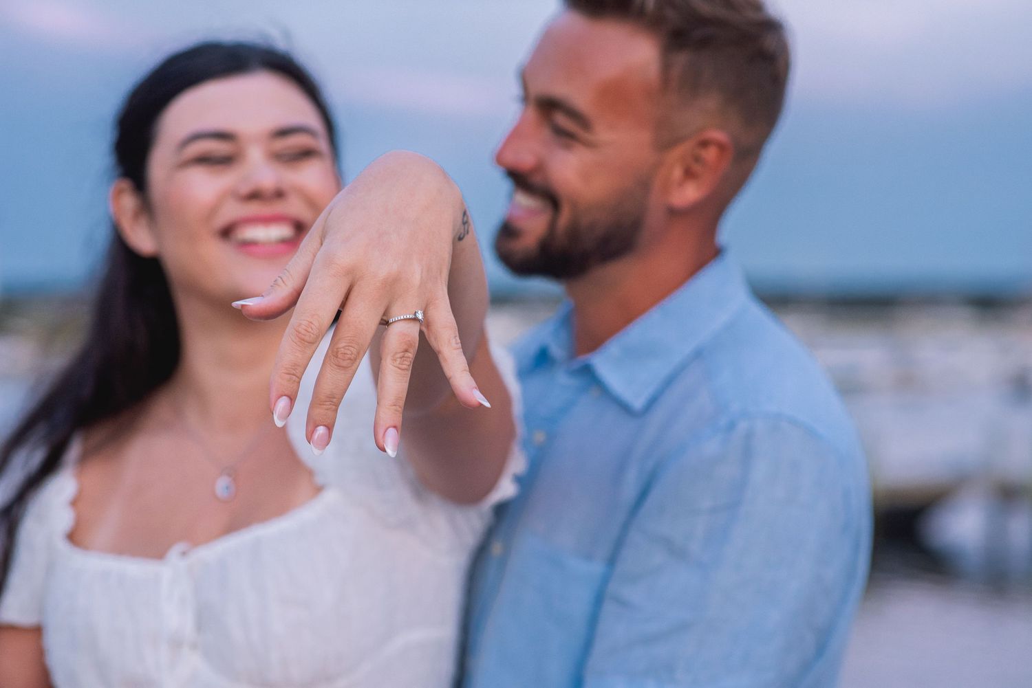 Happy couple showing off engagement ring outdoors at dusk, woman smiling and displaying diamond ring on her finger.