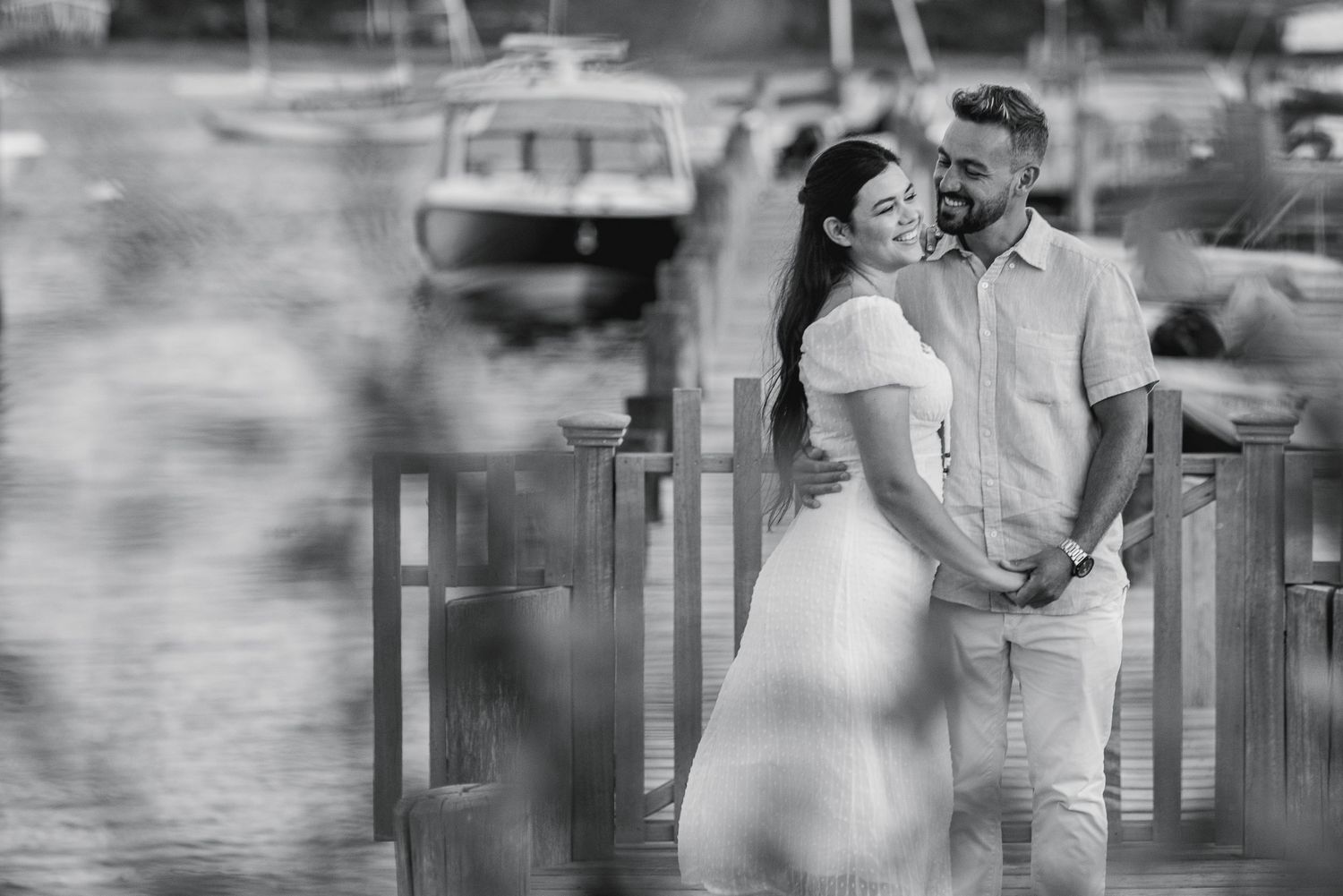 Black and white photo of a couple smiling and embracing on a dock near a marina waterfront.