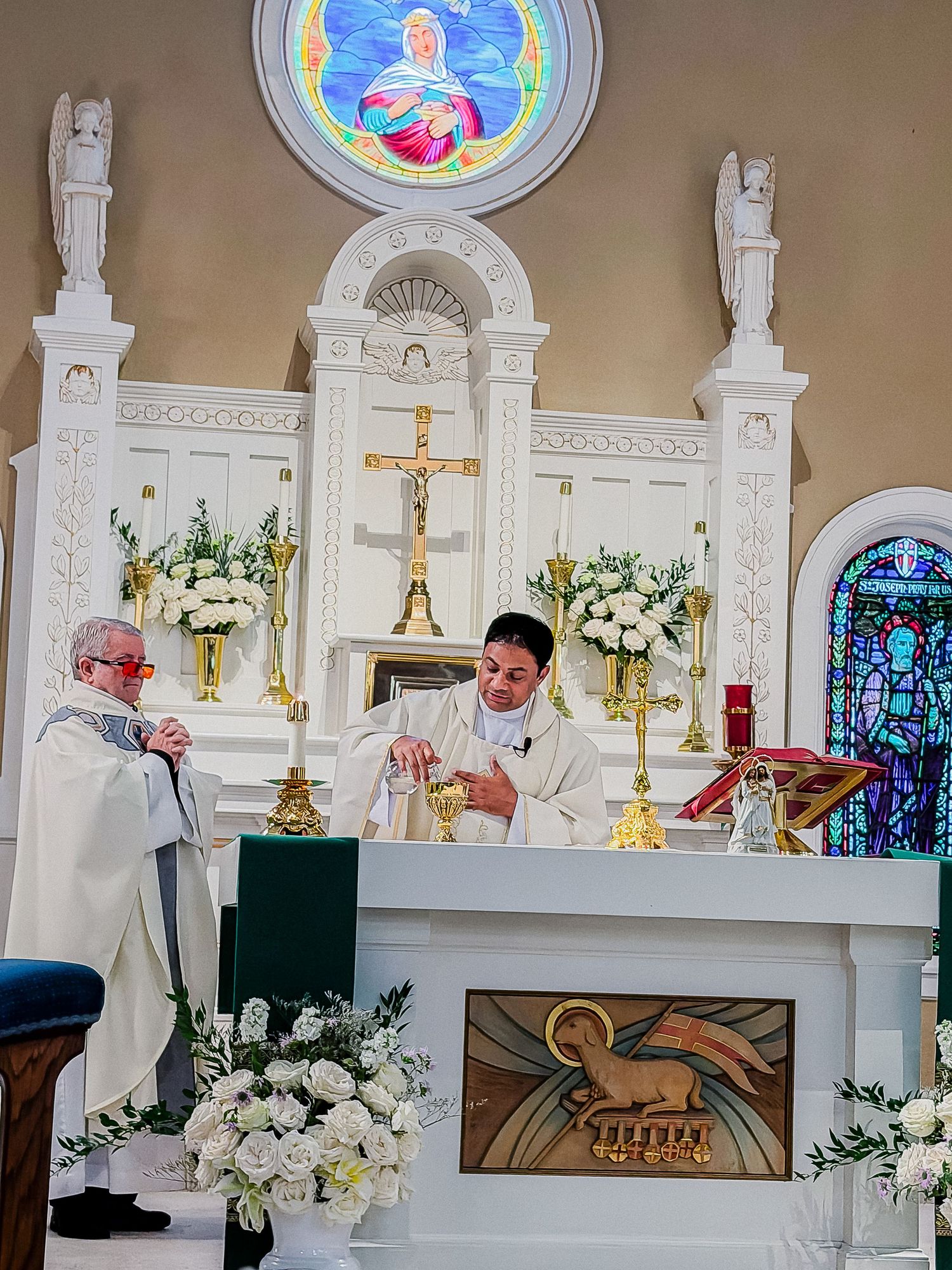 Catholic priests performing wedding Mass at ornate white altar adorned with white floral arrangements.