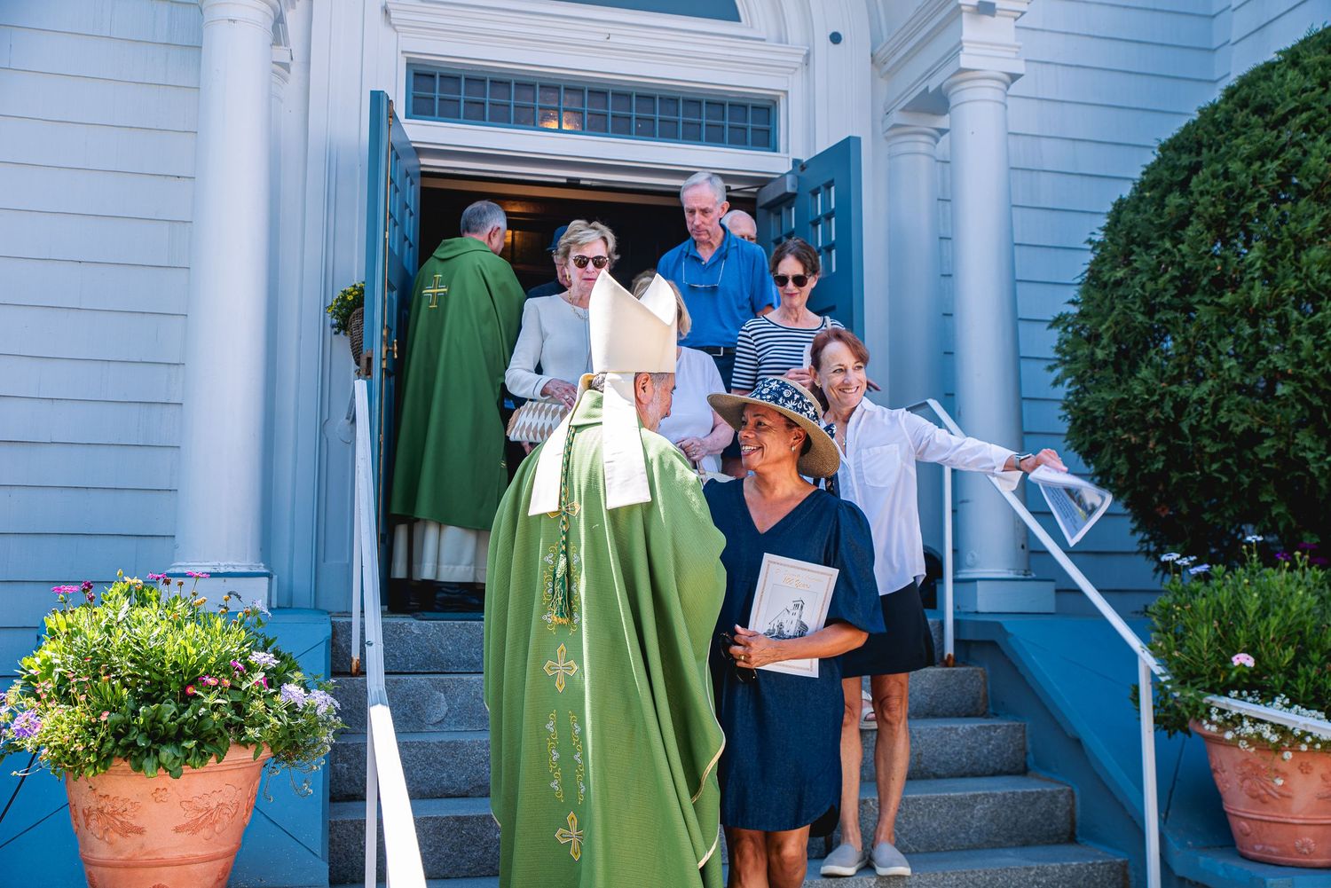 Priest in green vestments greeting parishioners on steps of a white church after Sunday service.