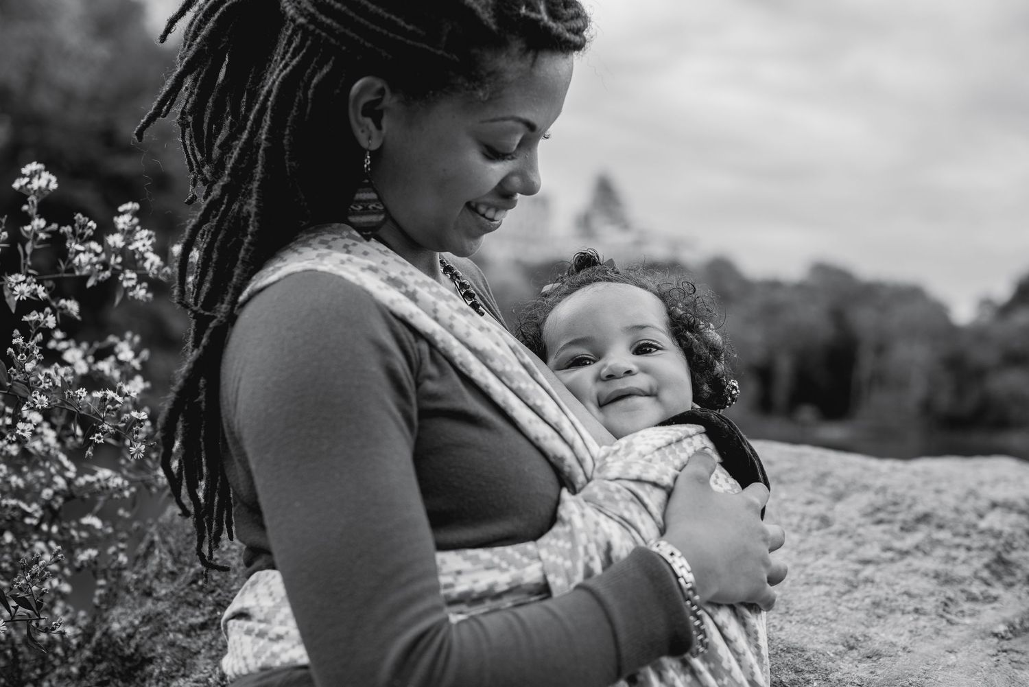 Black and white photo of a smiling mother with dreadlocks holding her happy baby in a wrap carrier outdoors.