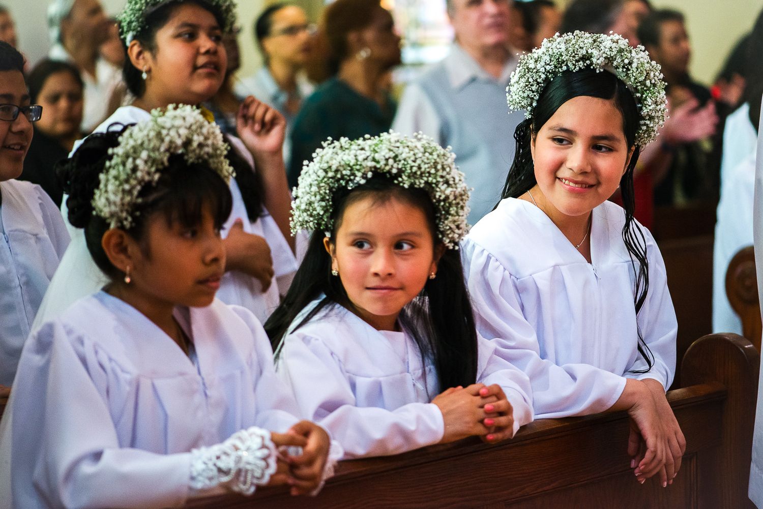 Three young girls in white dresses and floral crowns smile during a First Communion ceremony in a church.