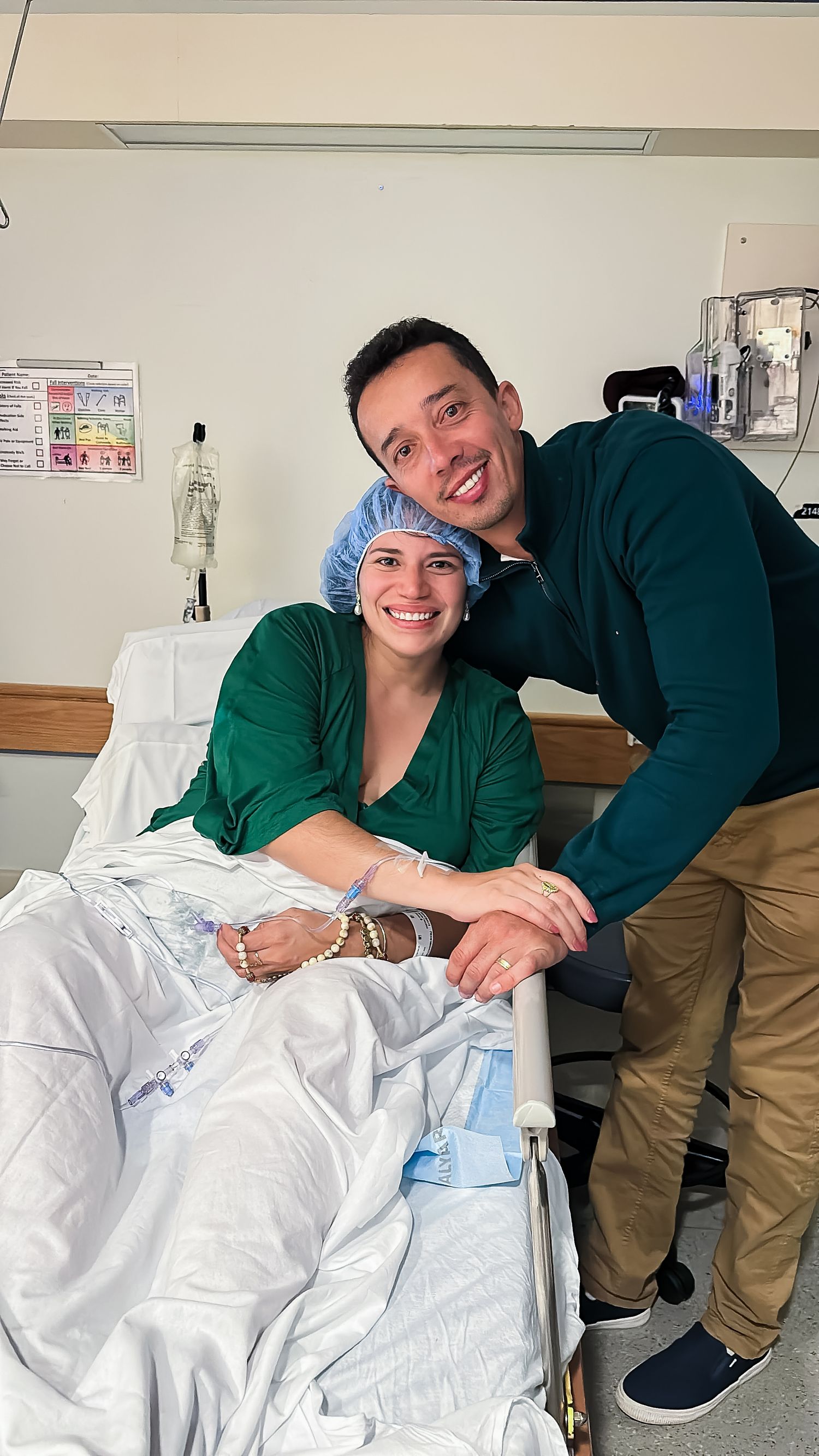 Smiling woman in a hospital bed wearing a blue surgical cap and green gown, with a smiling man in a green sweater leaning in beside her.