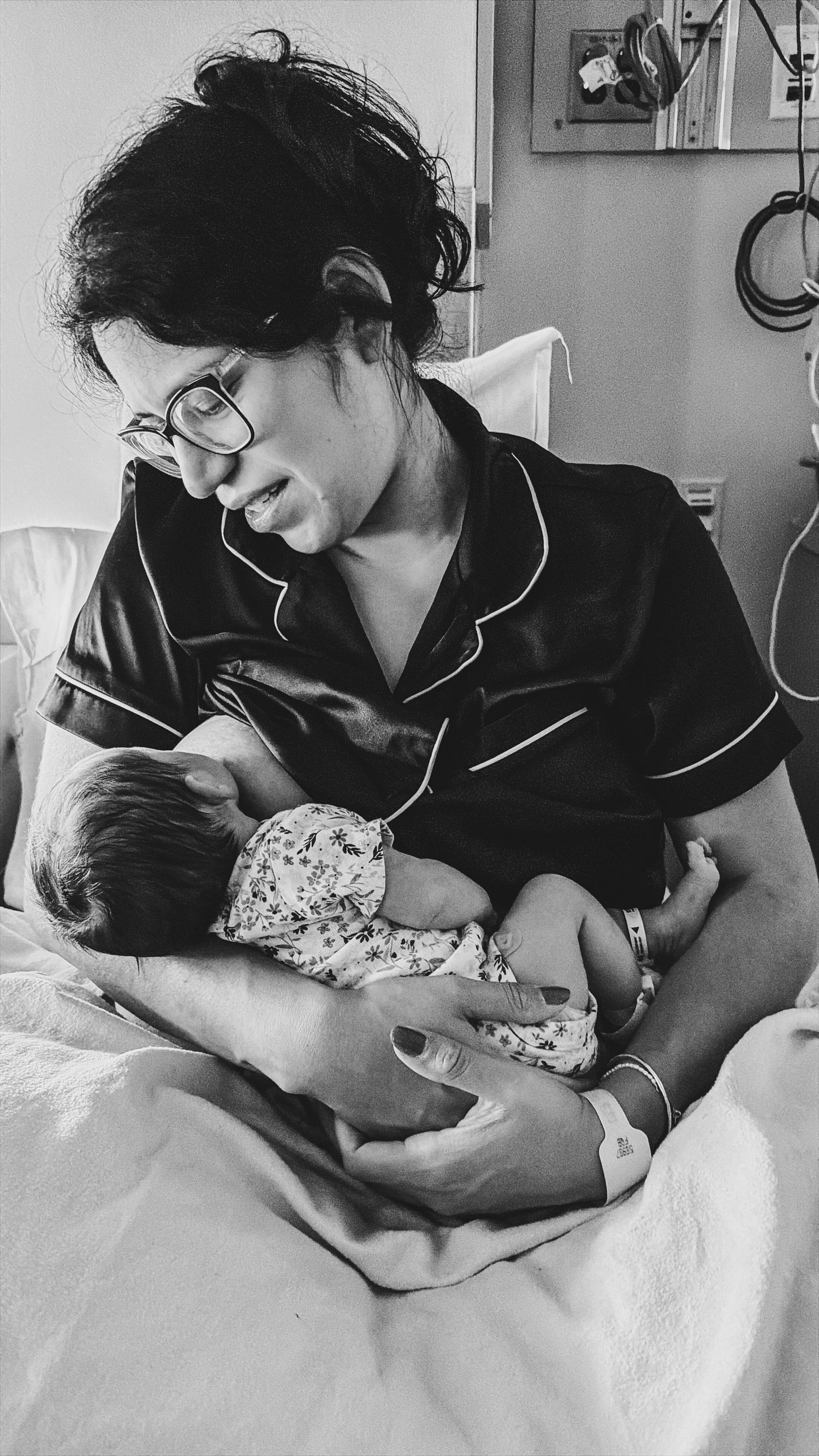 Black and white photo of a new mother in pajamas and glasses tenderly holding her newborn baby in a hospital bed.
