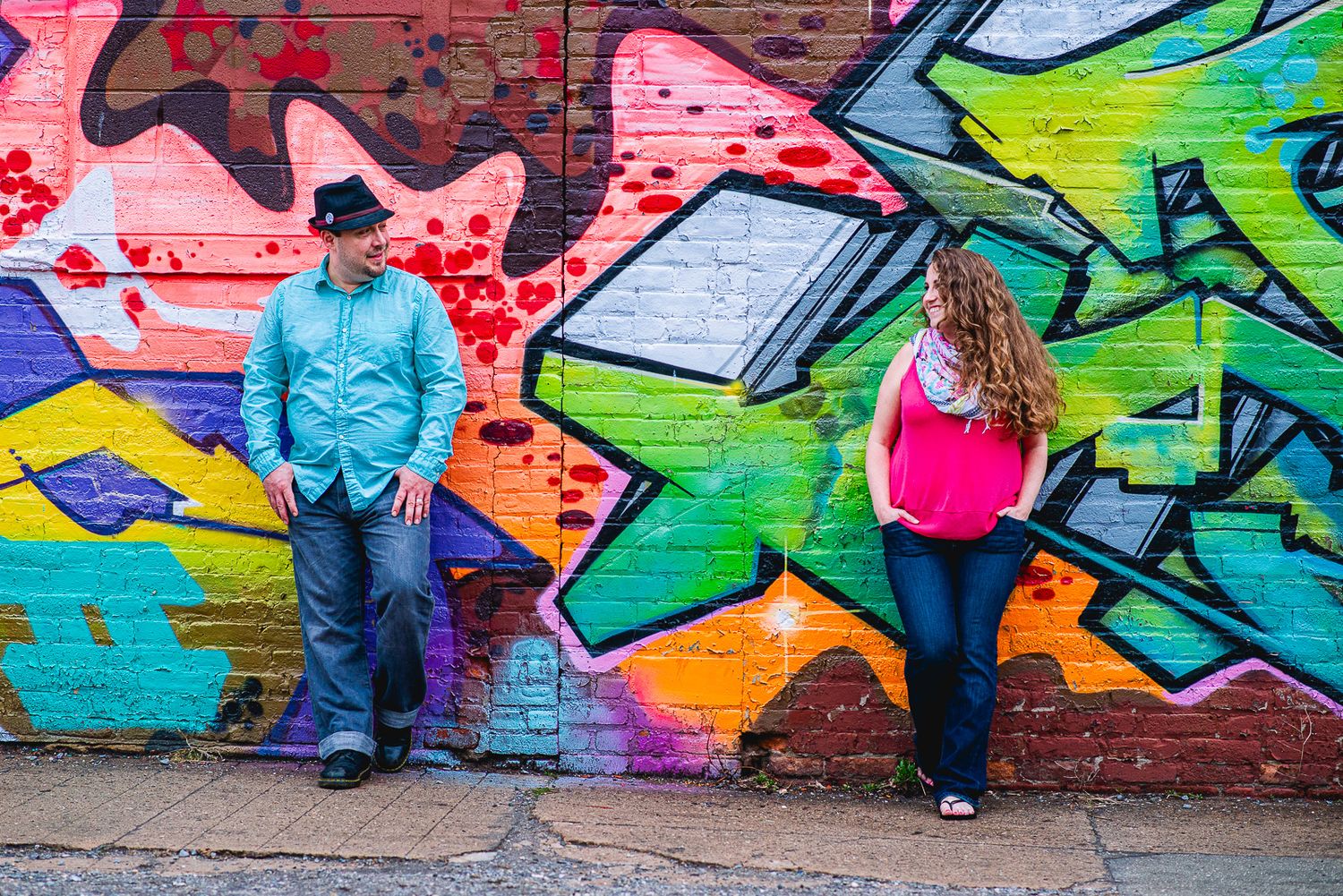 Two people stand apart against a vibrant colorful graffiti brick wall, one in a blue shirt and hat, other in a pink top.