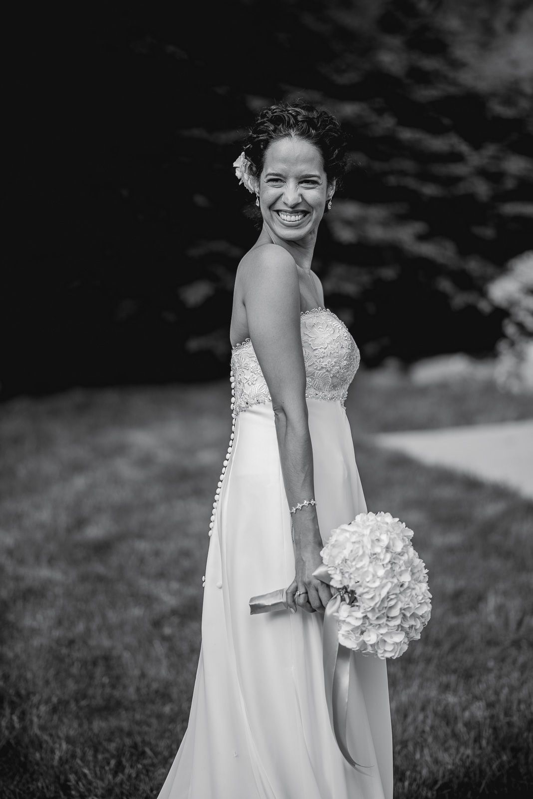 Smiling bride in strapless lace gown holding hydrangea bouquet, black and white outdoor wedding portrait.