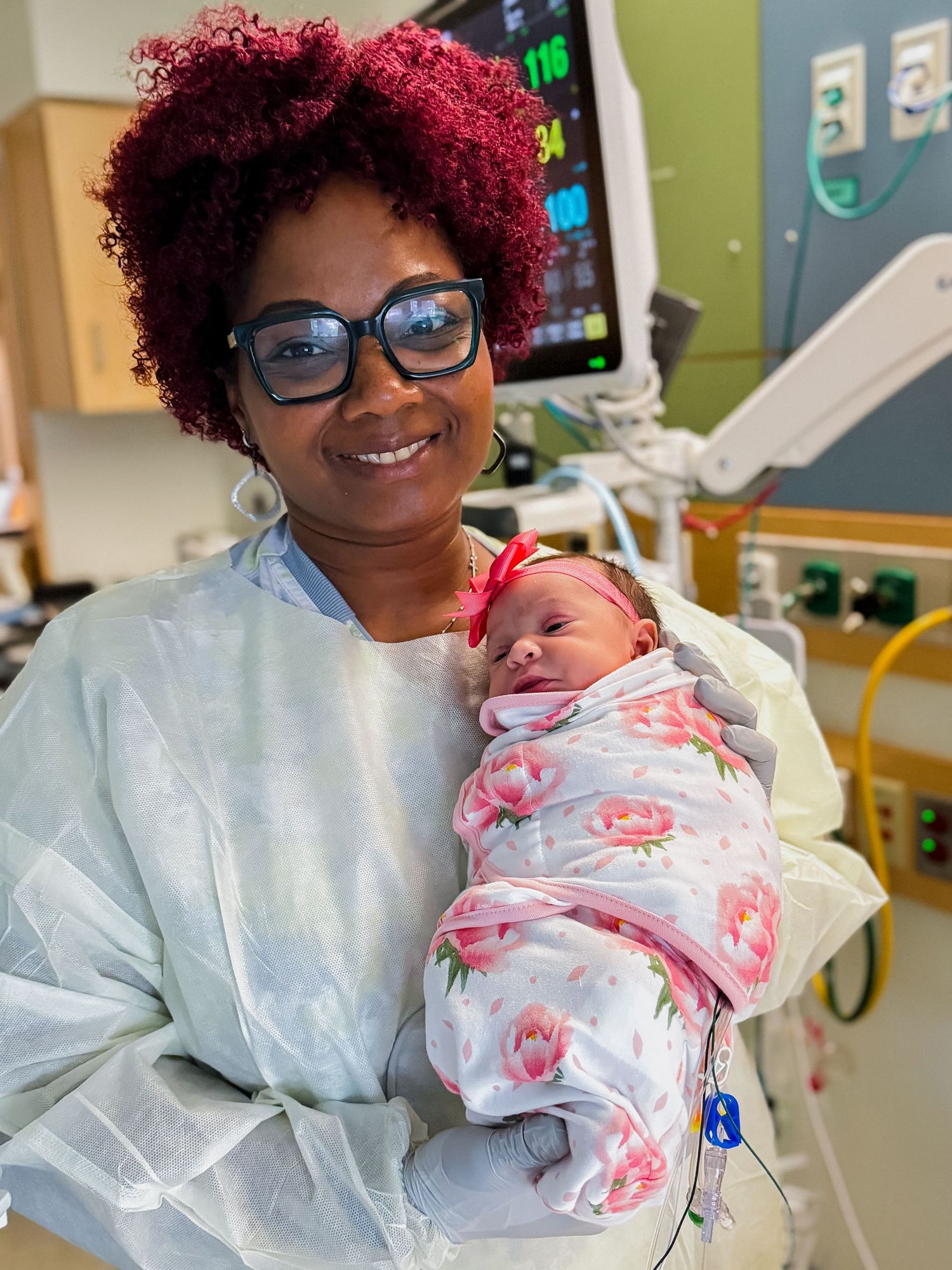 Smiling woman in glasses and a protective gown holds a newborn in a floral swaddle and pink headband in a hospital room with monitors.