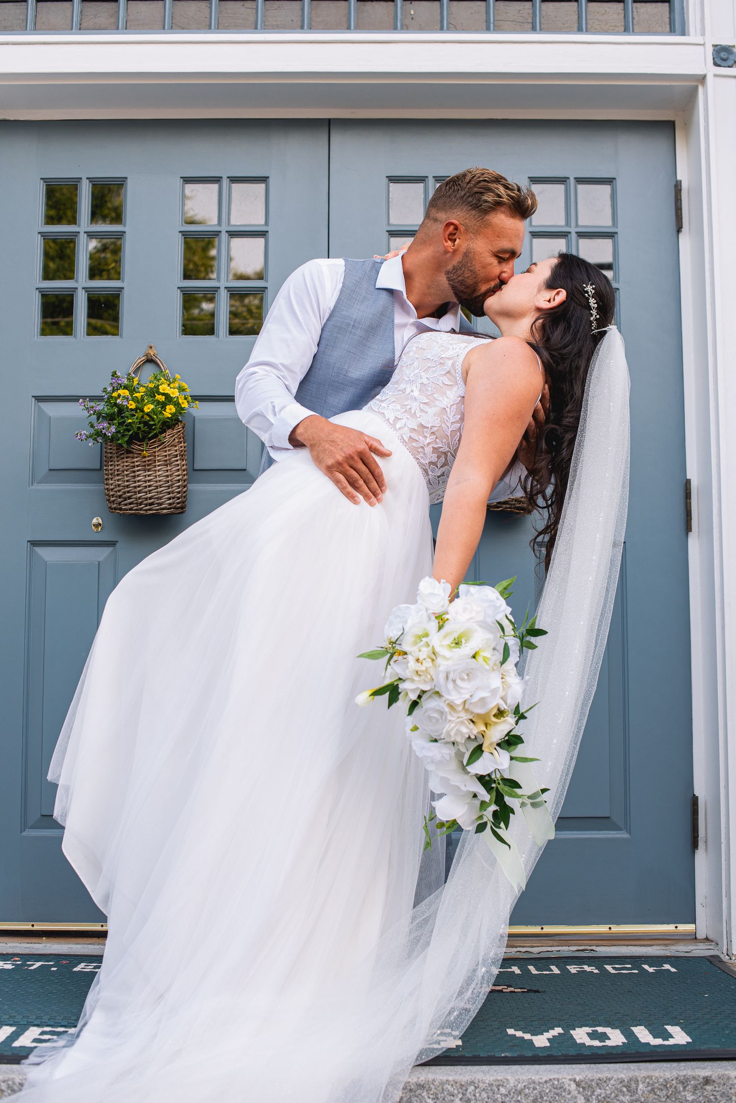 Bride and groom share a romantic kiss outside blue church doors, bride holding white floral bouquet.