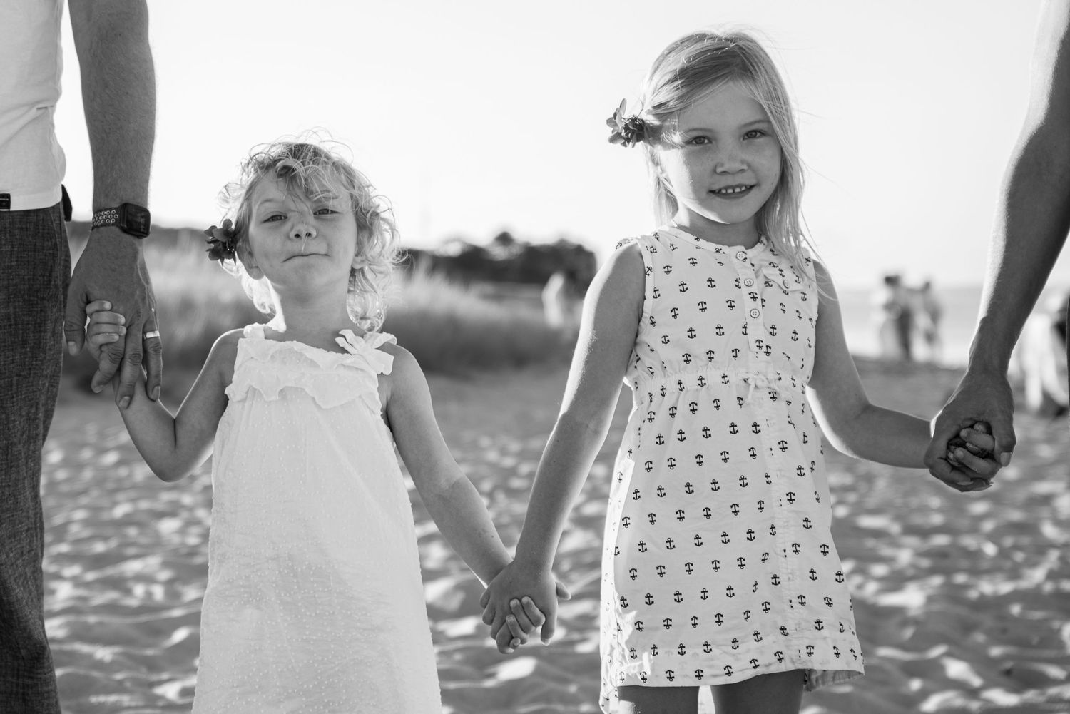 Black and white photo of two little girls in dresses holding hands on a sandy beach.