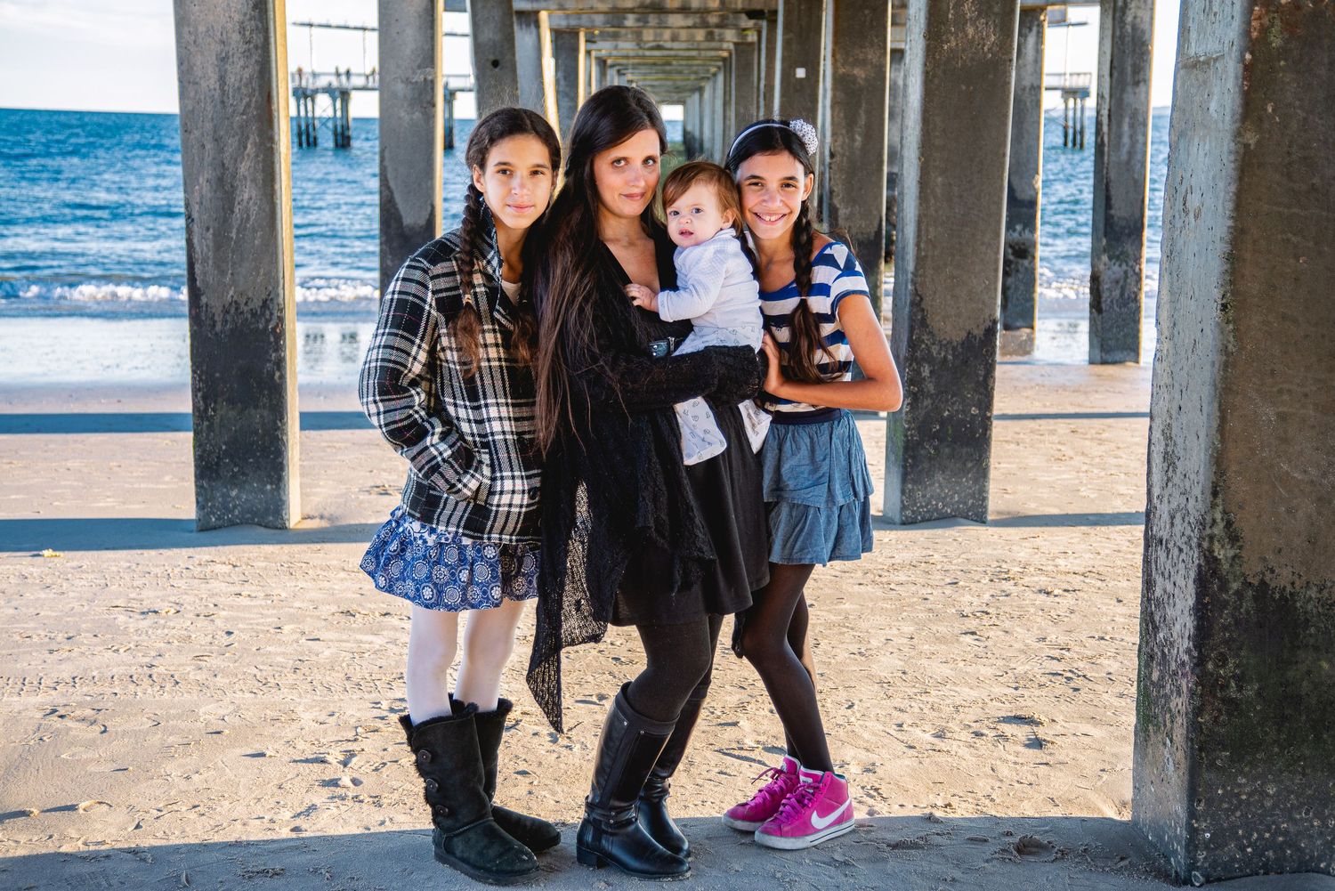 A woman holds a baby while posing with two girls under a beach pier on a cloudy day.