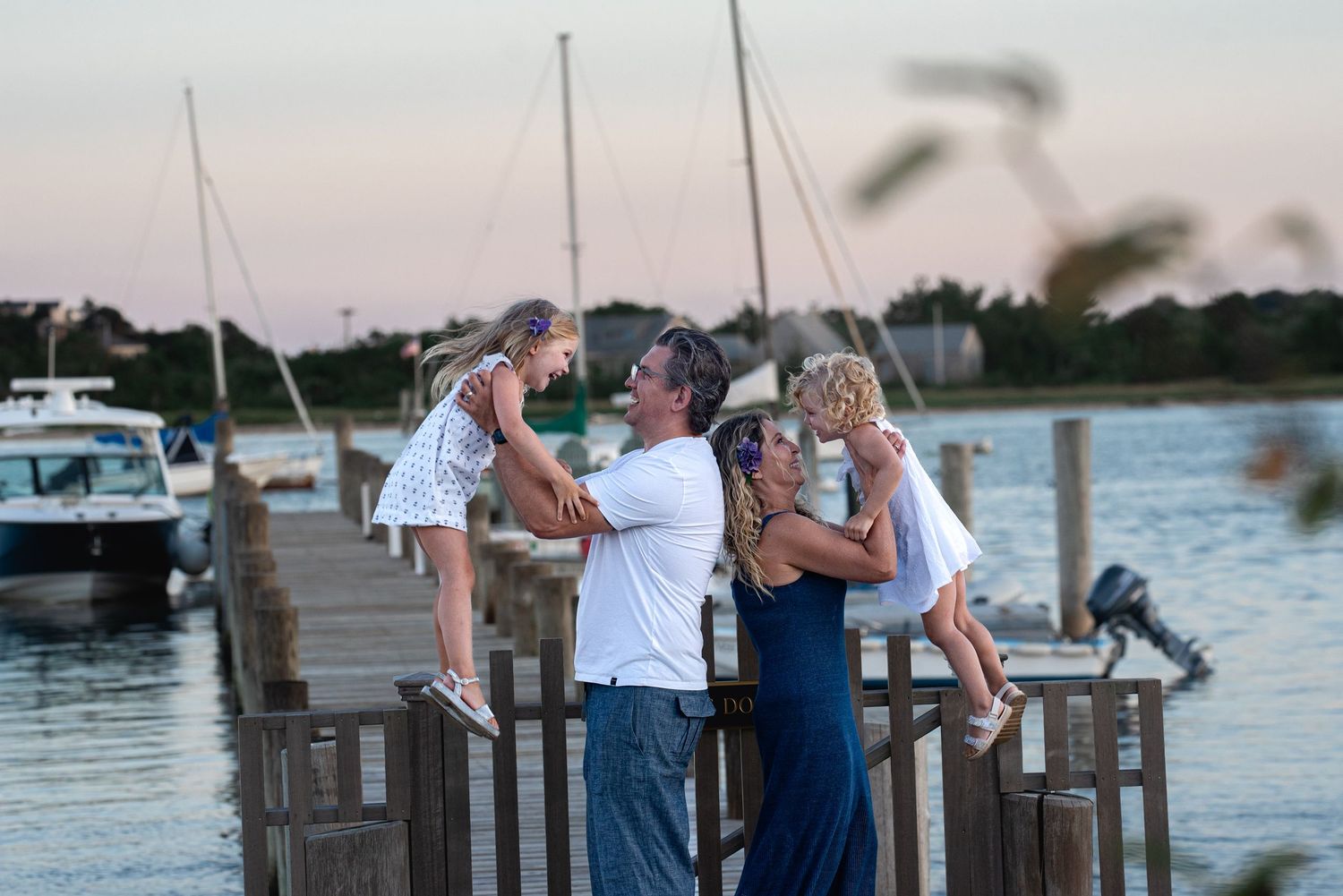 Family of four enjoying a sunset at a marina dock, parents lifting two young girls in white dresses near sailboats.