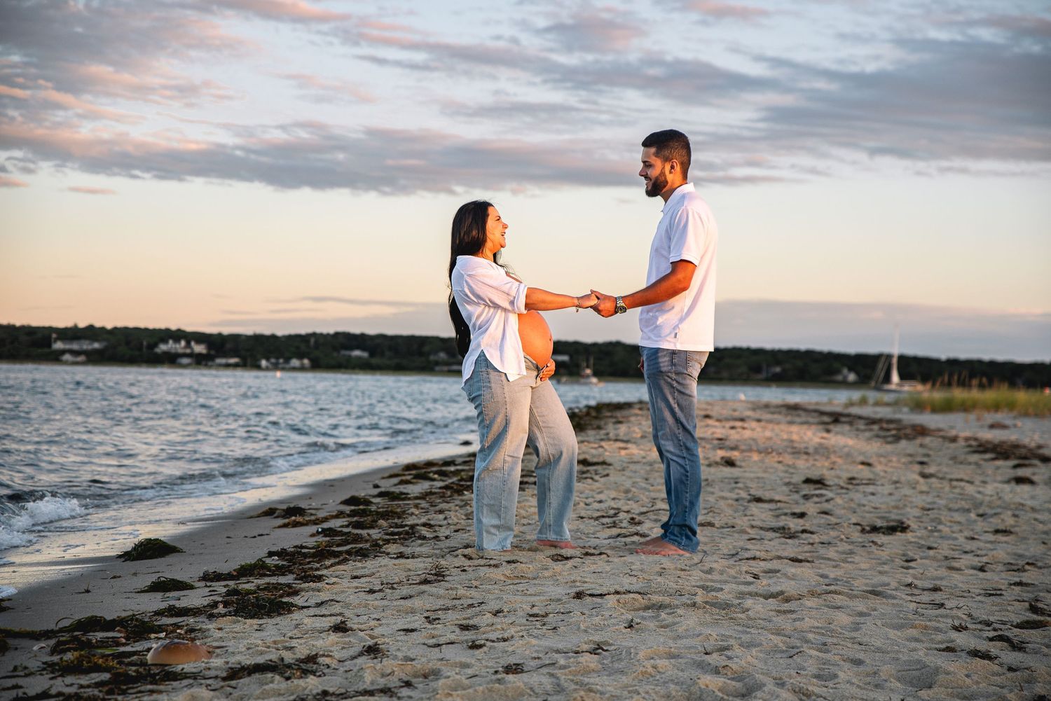 A couple holds hands on a sandy beach at sunset, wearing white shirts and jeans with ocean waves behind them.
