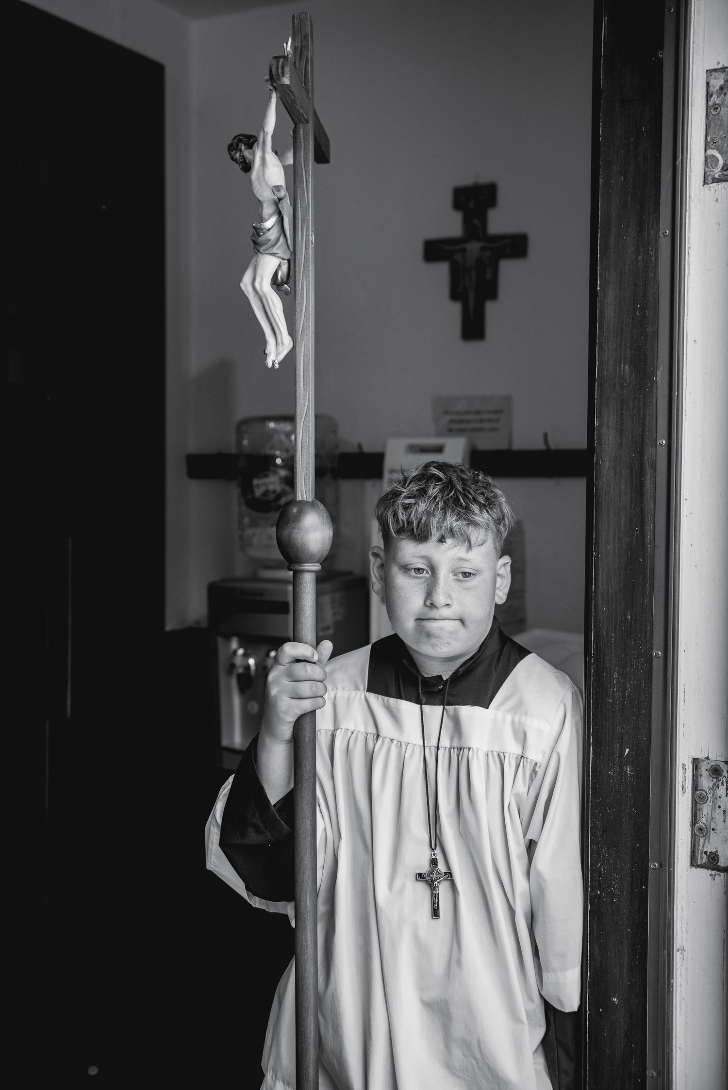 Young altar boy in white vestments holds a crucifix staff, with a cross visible on the wall behind him.